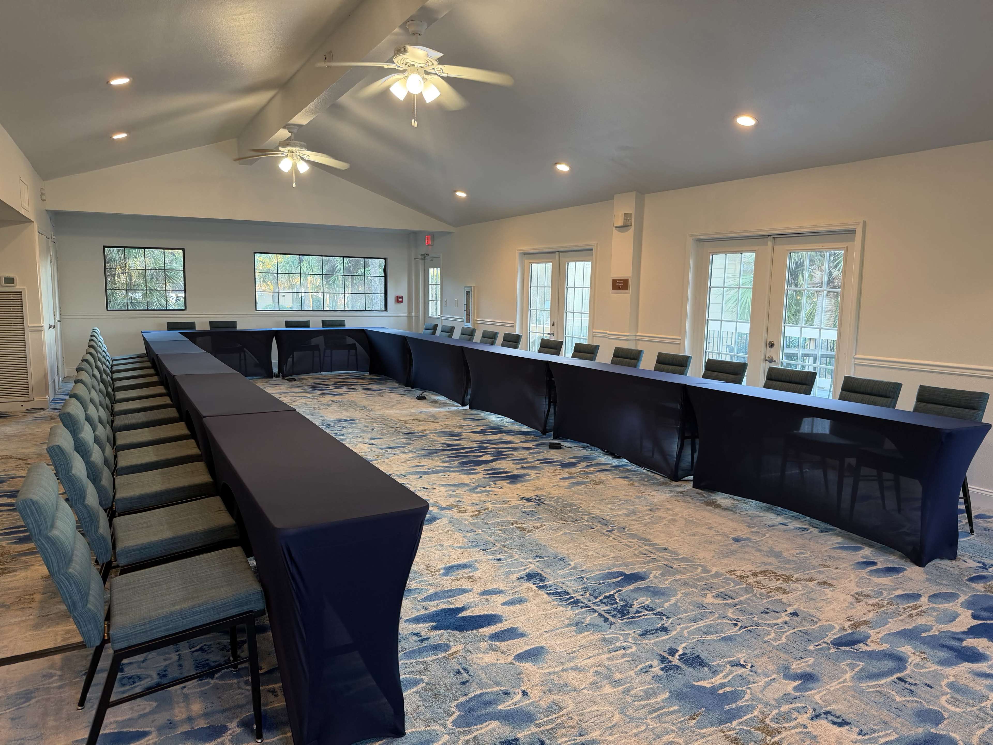 A large room features a U-shaped arrangement of tables covered with navy tablecloths, surrounded by chairs, in a well-lit space with large windows.