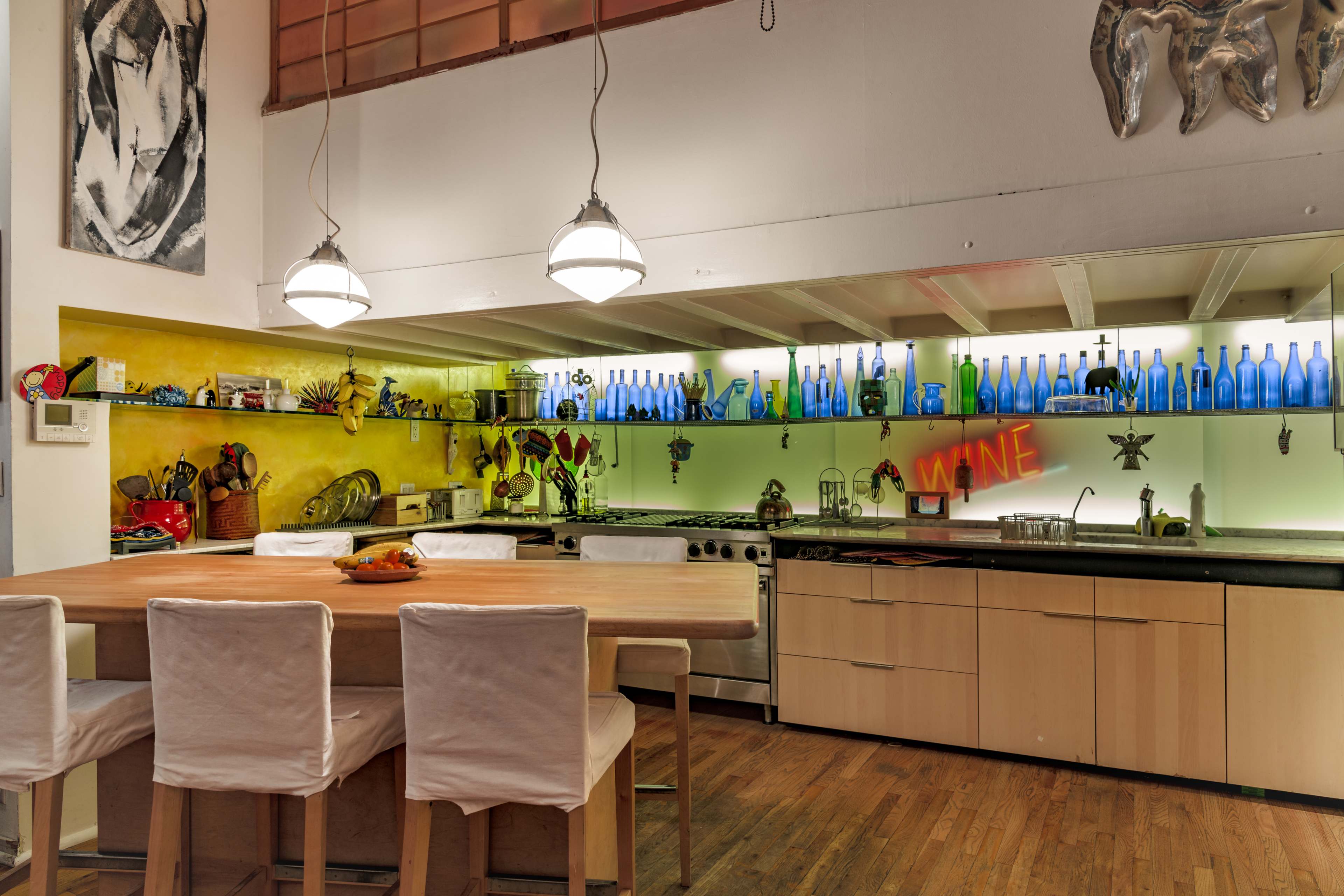 A modern kitchen with a large wooden dining table, illuminated shelves containing blue glass bottles, and various kitchen utensils displayed along the countertop.
