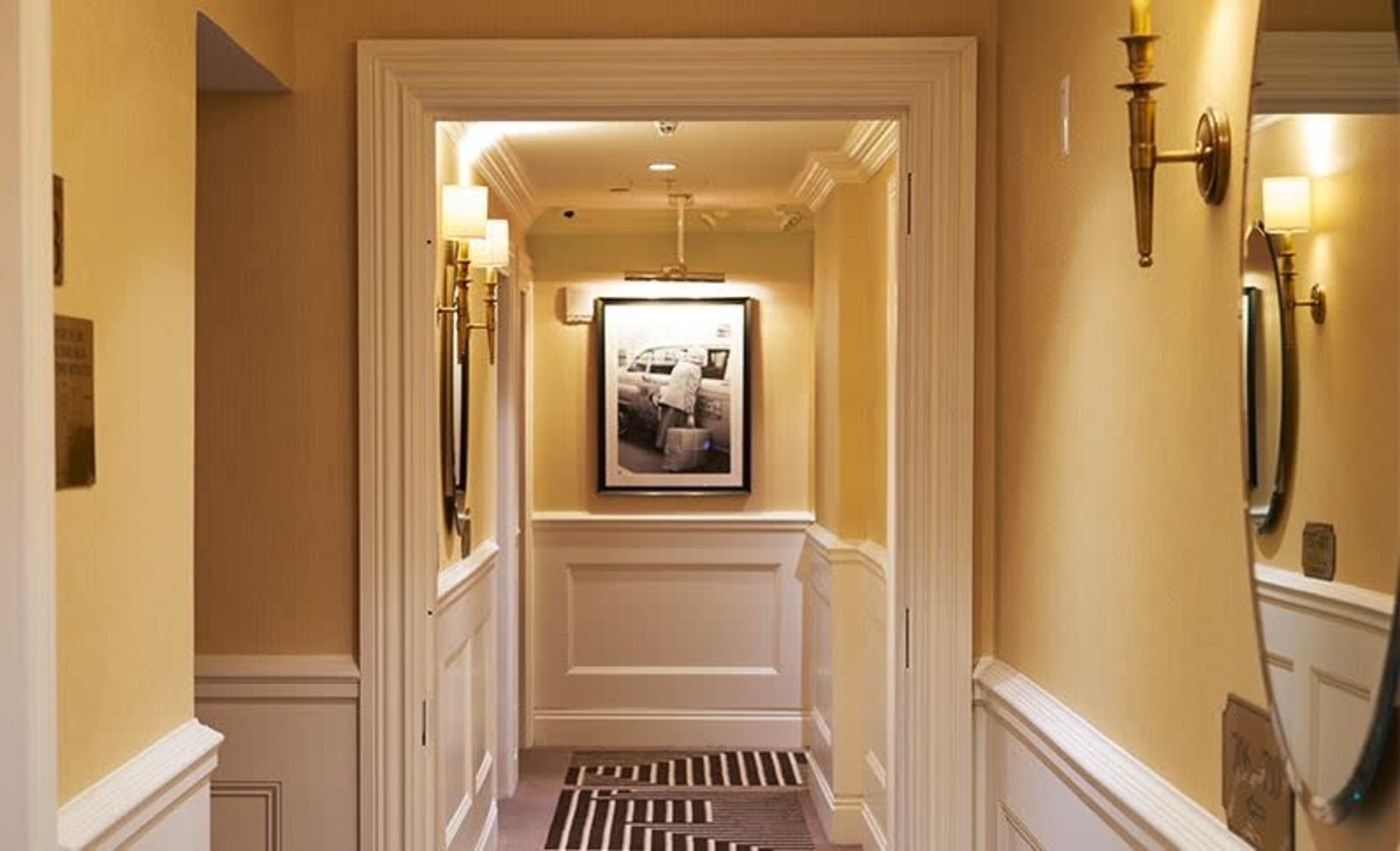 A well-lit hotel hallway featuring a framed photograph on the wall and sconces on either side.