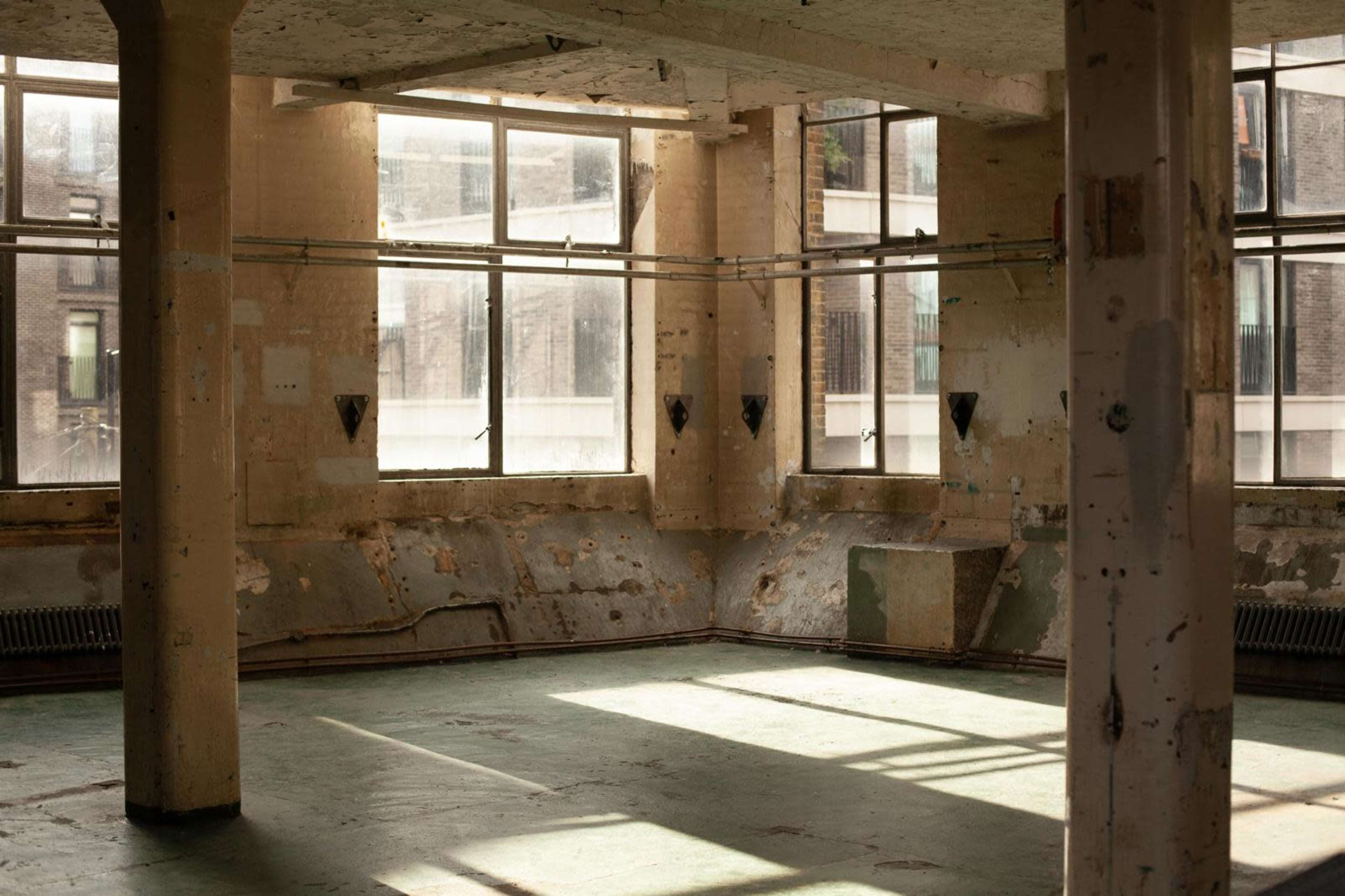 The image shows an empty, industrial room with large windows allowing light to enter, highlighting the worn walls and floor.