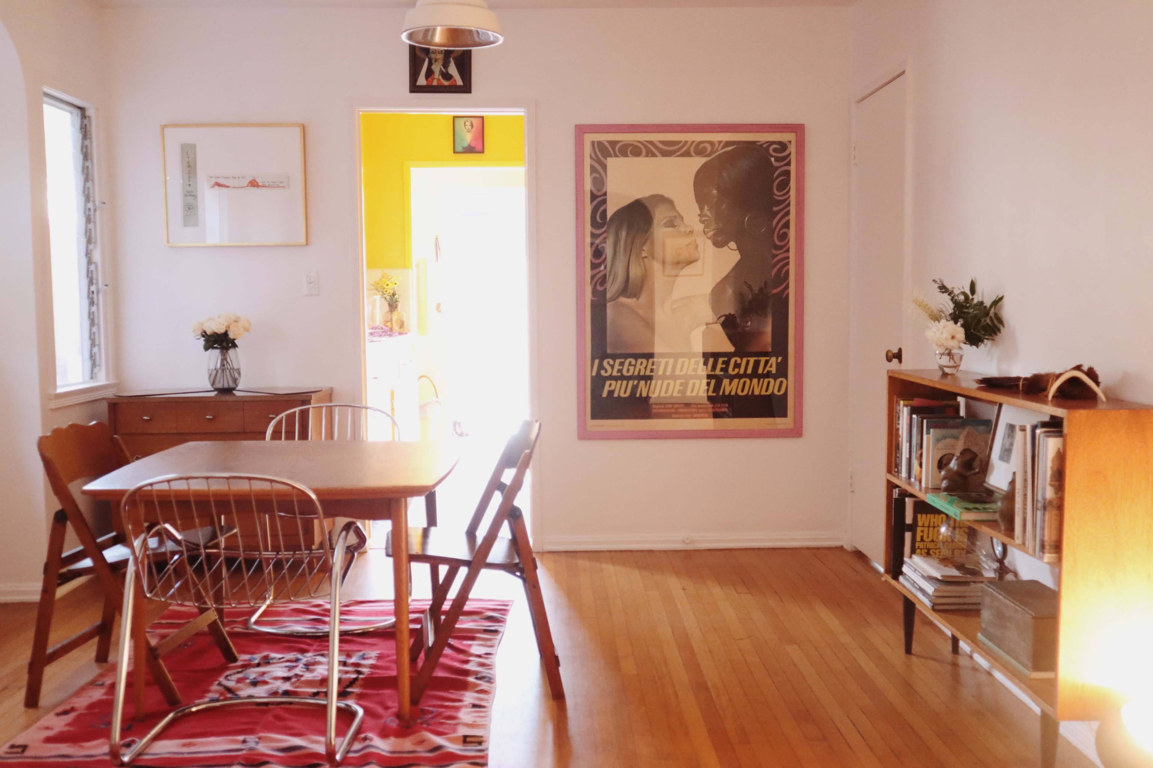 A cozy room featuring a wooden dining table surrounded by chairs, complemented by a large poster on the wall and a bookshelf nearby.