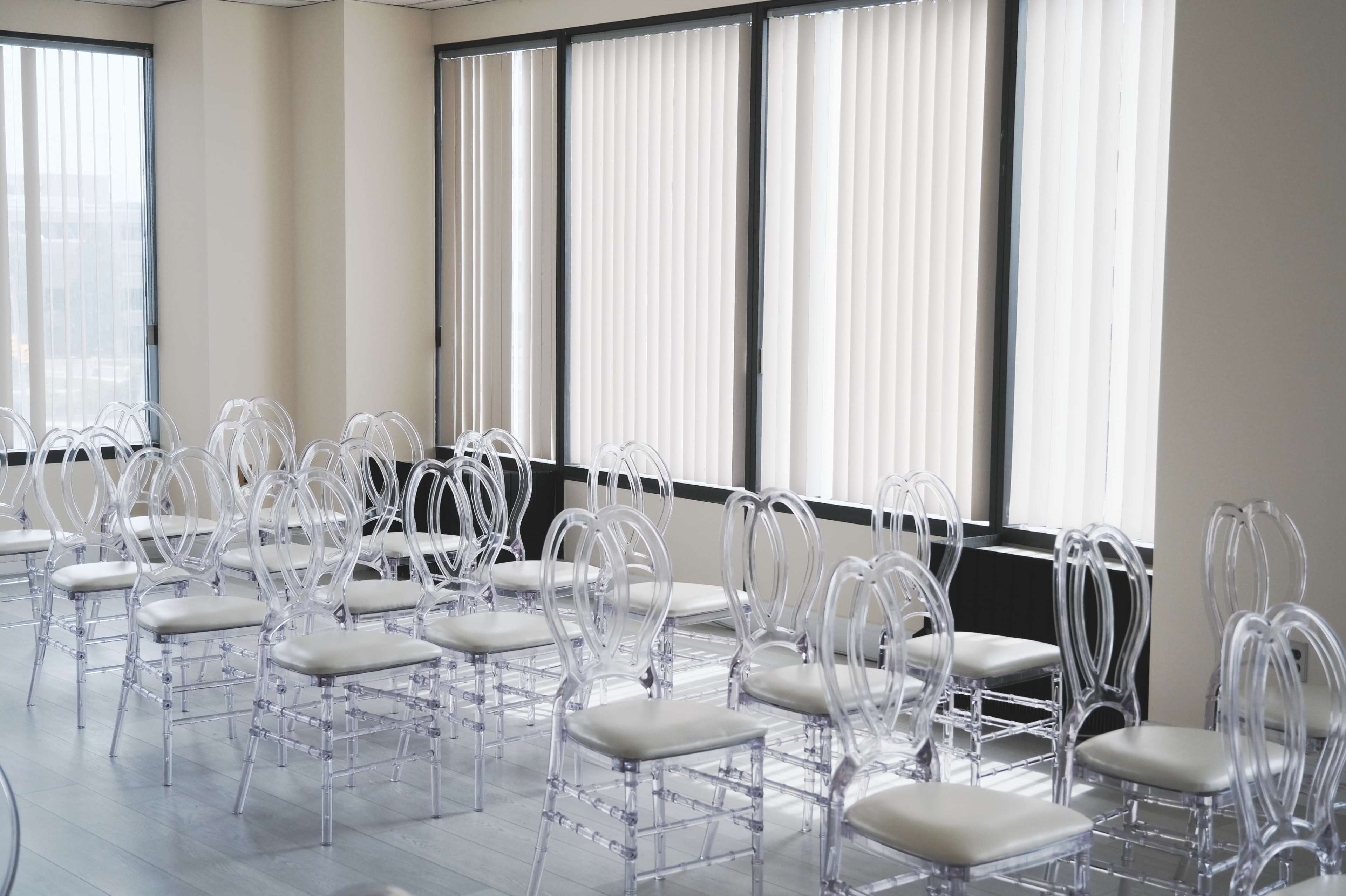 A row of clear plastic chairs is arranged in a well-lit room with large windows covered by vertical blinds.