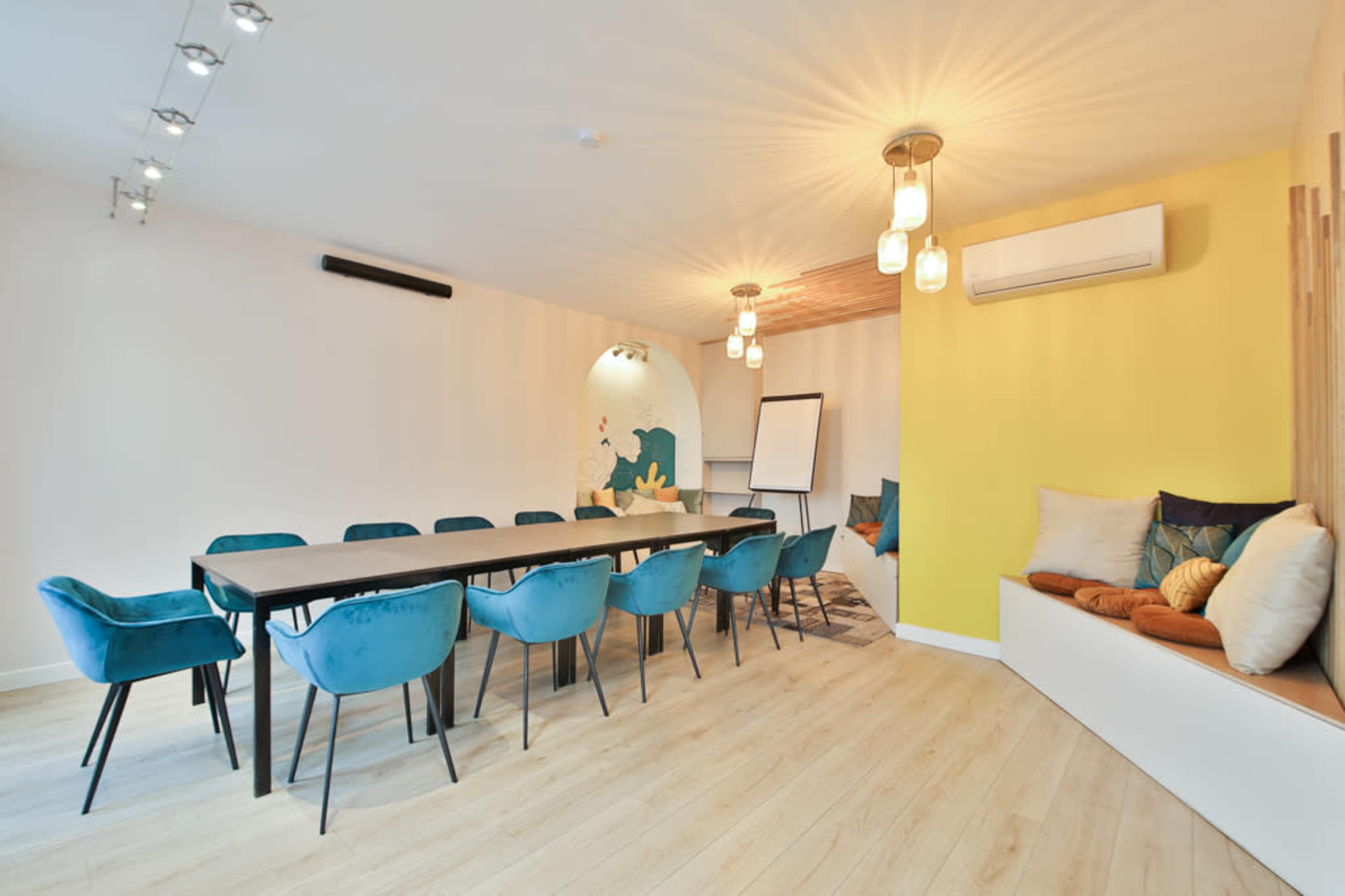 A modern meeting room features a long table surrounded by blue chairs, a yellow accent wall, and a cozy seating area with pillows.