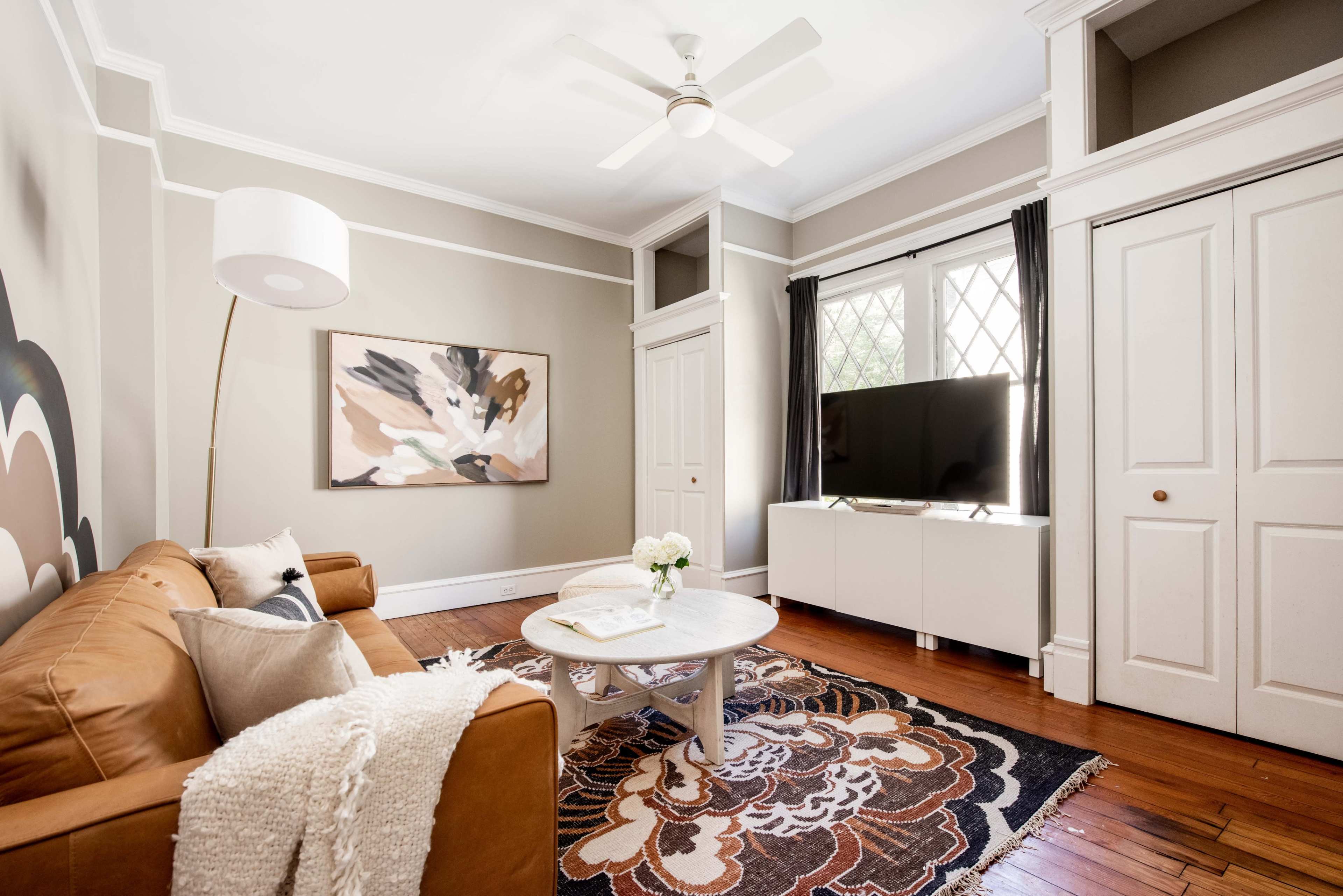 The living room features a brown sofa, a round coffee table, a large television, and a decorative area rug, all arranged around a window with curtains.