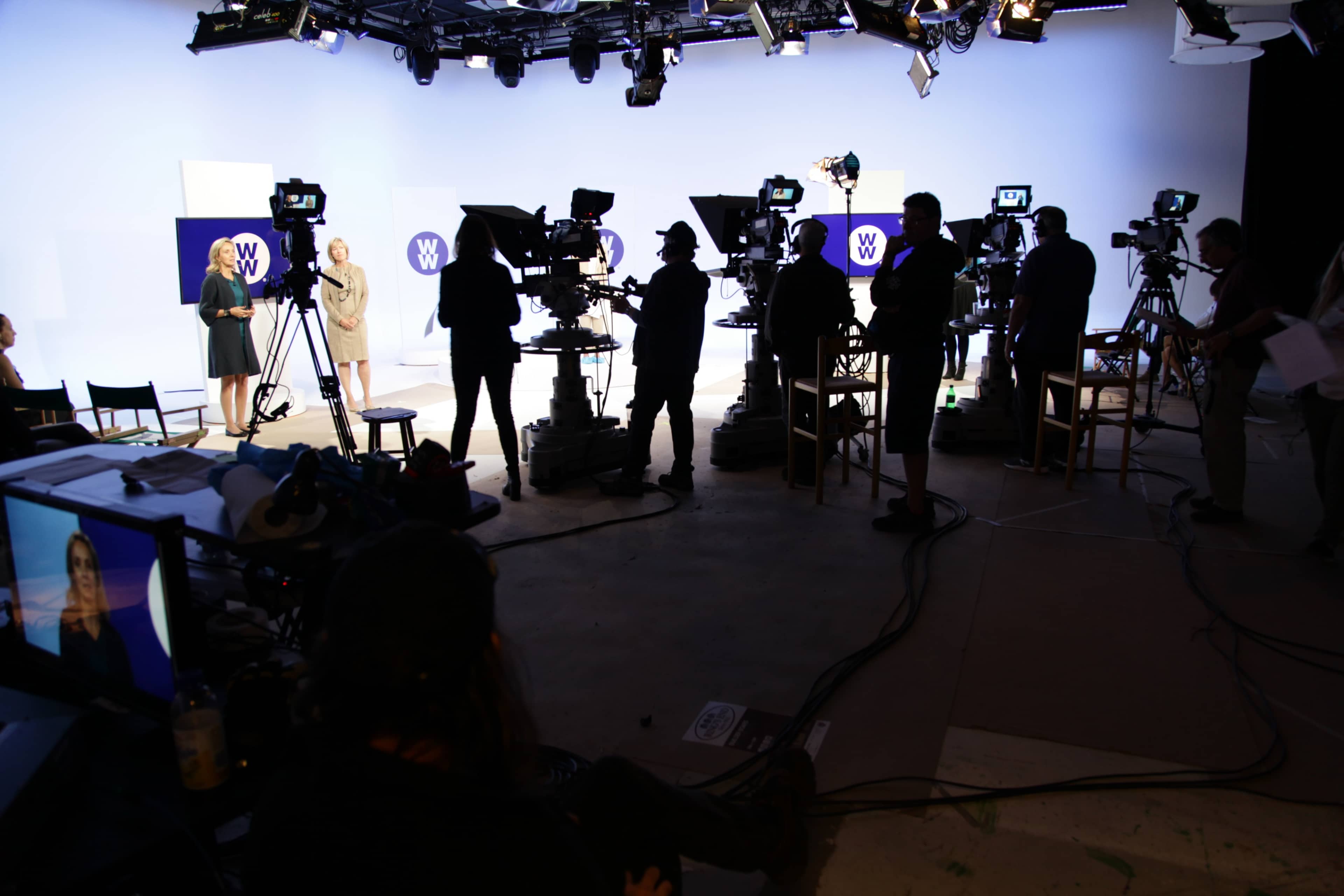 A film studio set with multiple cameras and crew members preparing for a shoot, while two women stand in front of a backdrop.