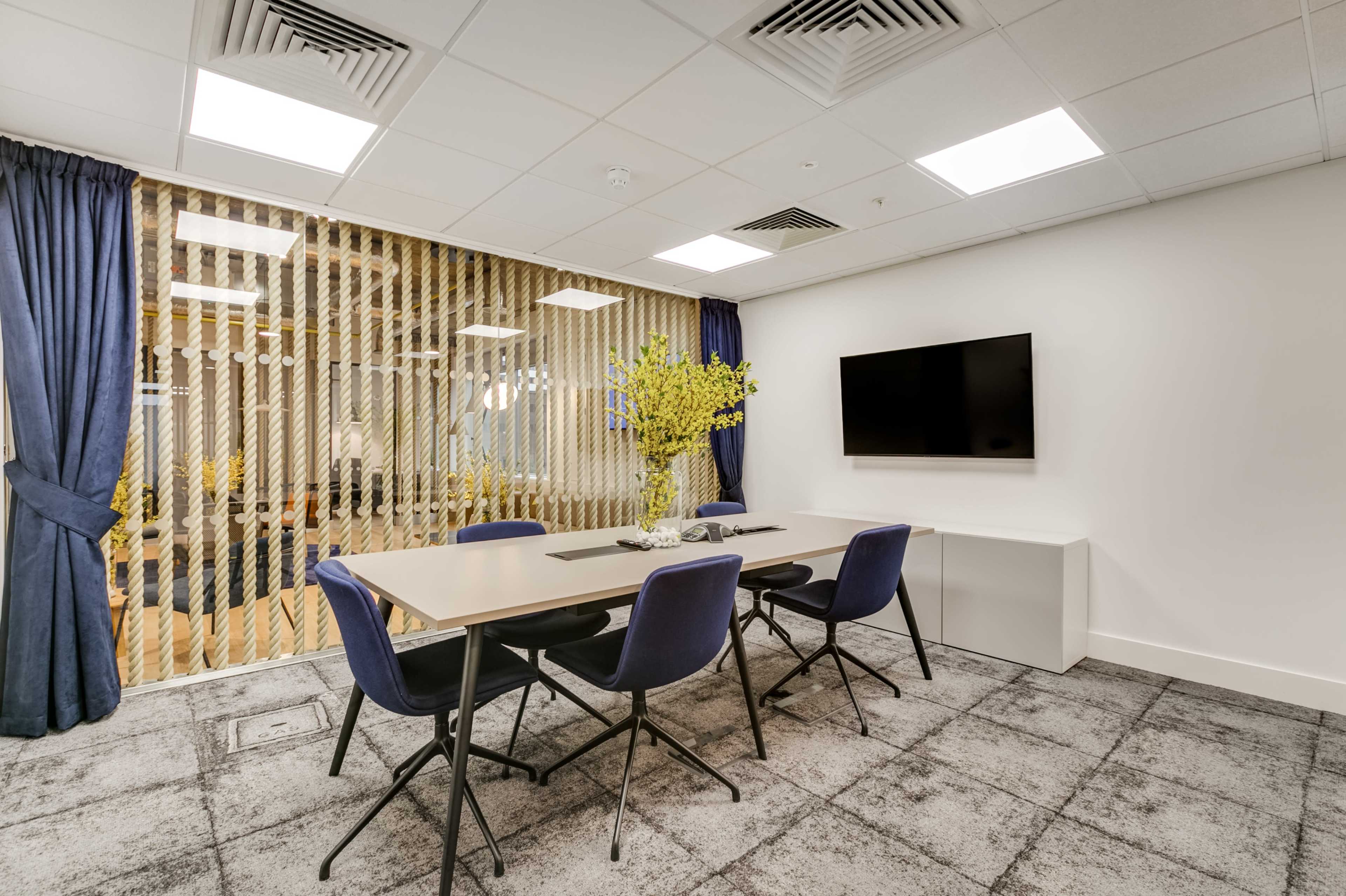A meeting room features a large table with four chairs, a wall-mounted TV, and a window covered with vertical blinds that offers a view of a light-filled space beyond.