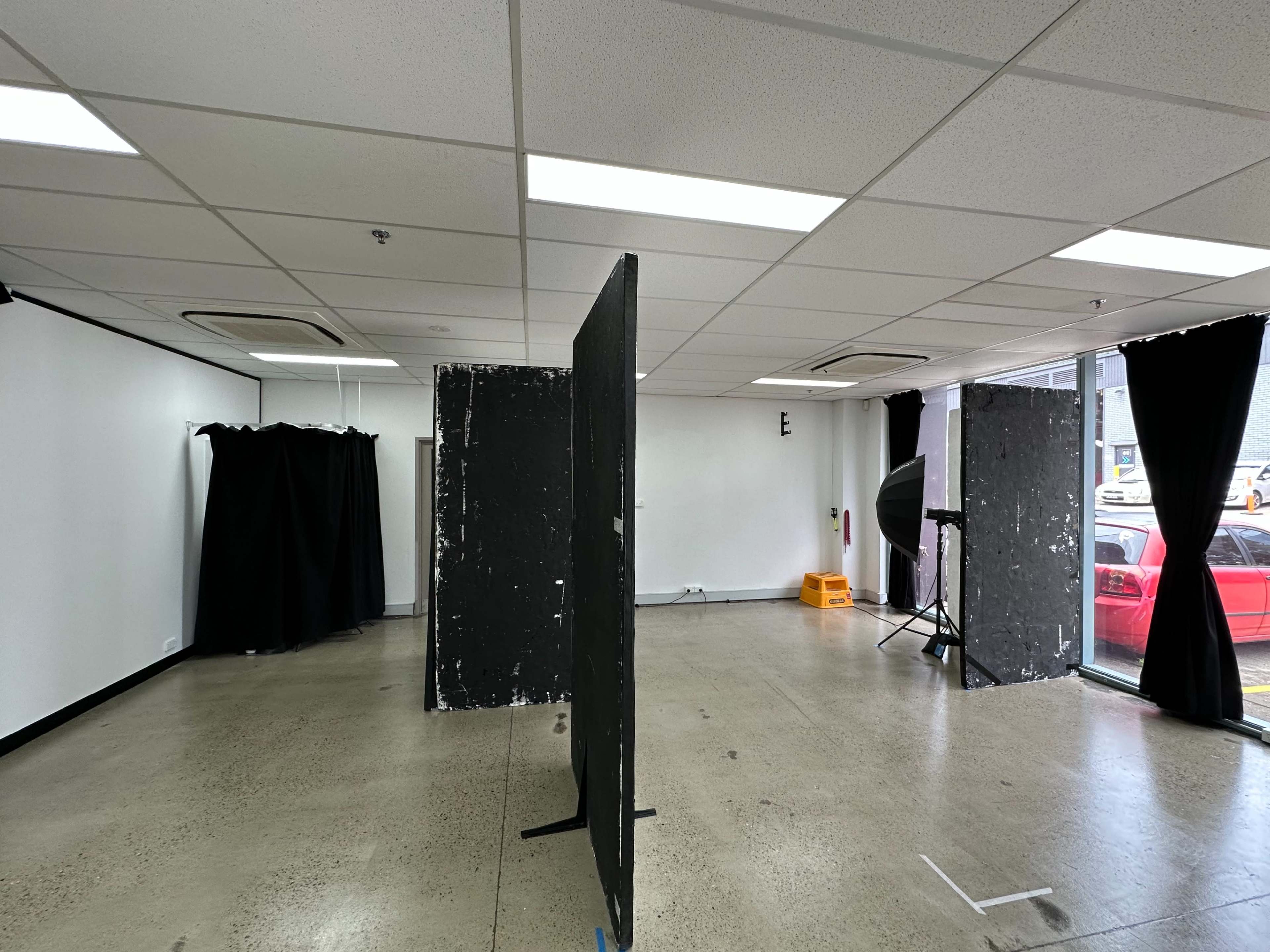 An empty room features black dividers, a photography light setup, and large windows revealing a red car outside.