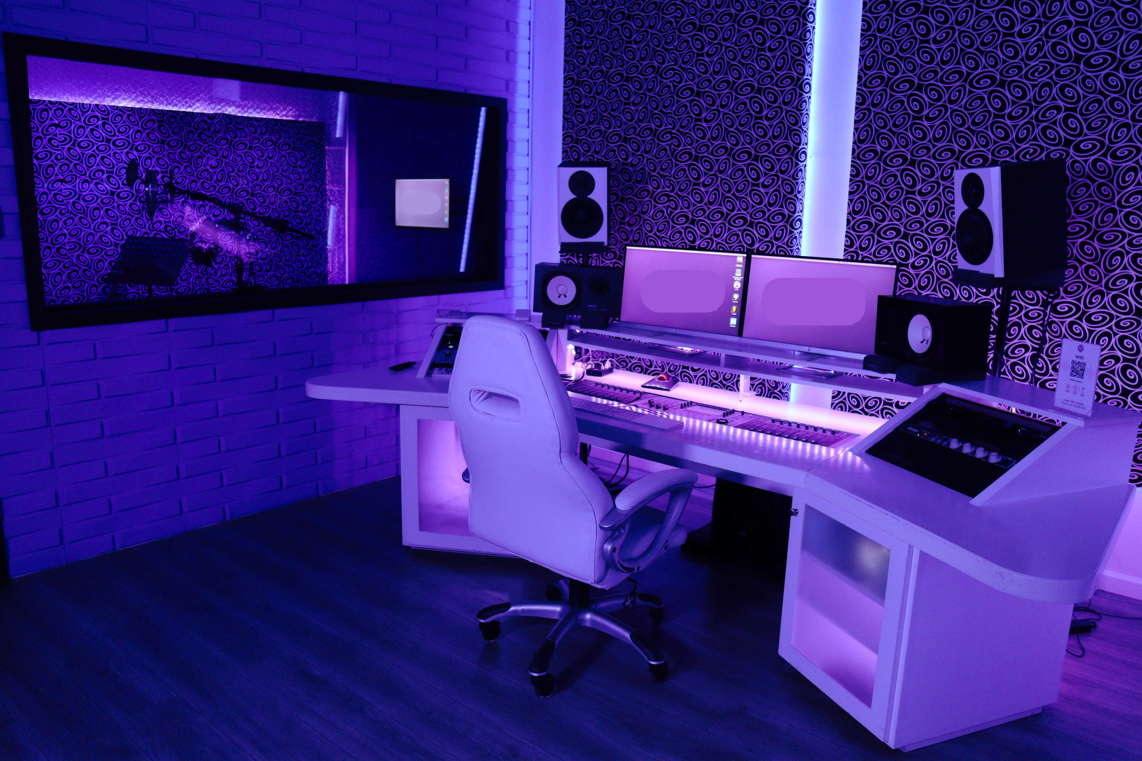 A modern music studio with a white desk, computer monitors, speakers, and purple lighting.