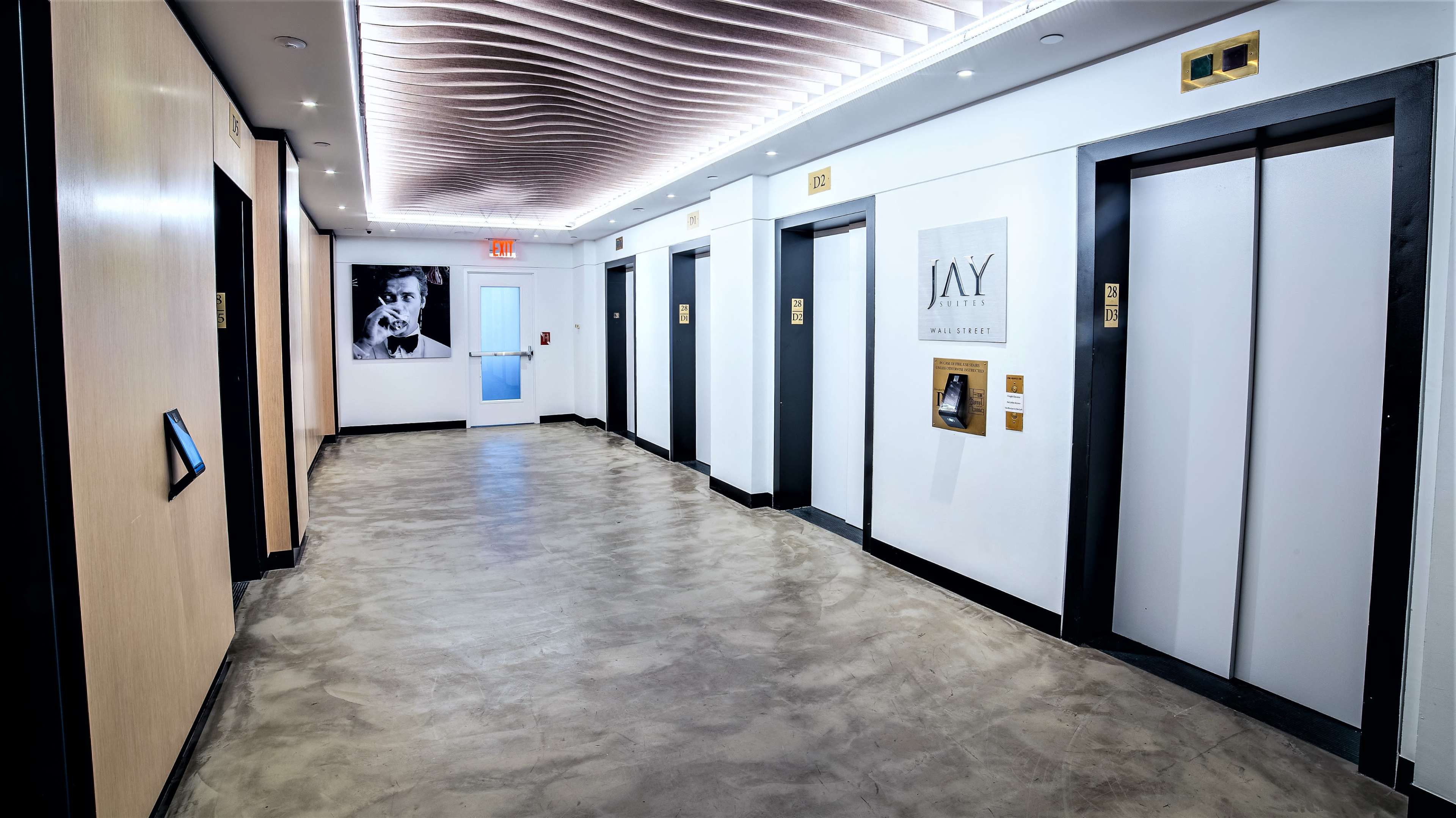 The image shows a corridor in a building featuring several elevator doors, a framed portrait on the wall, and a door at the far end.