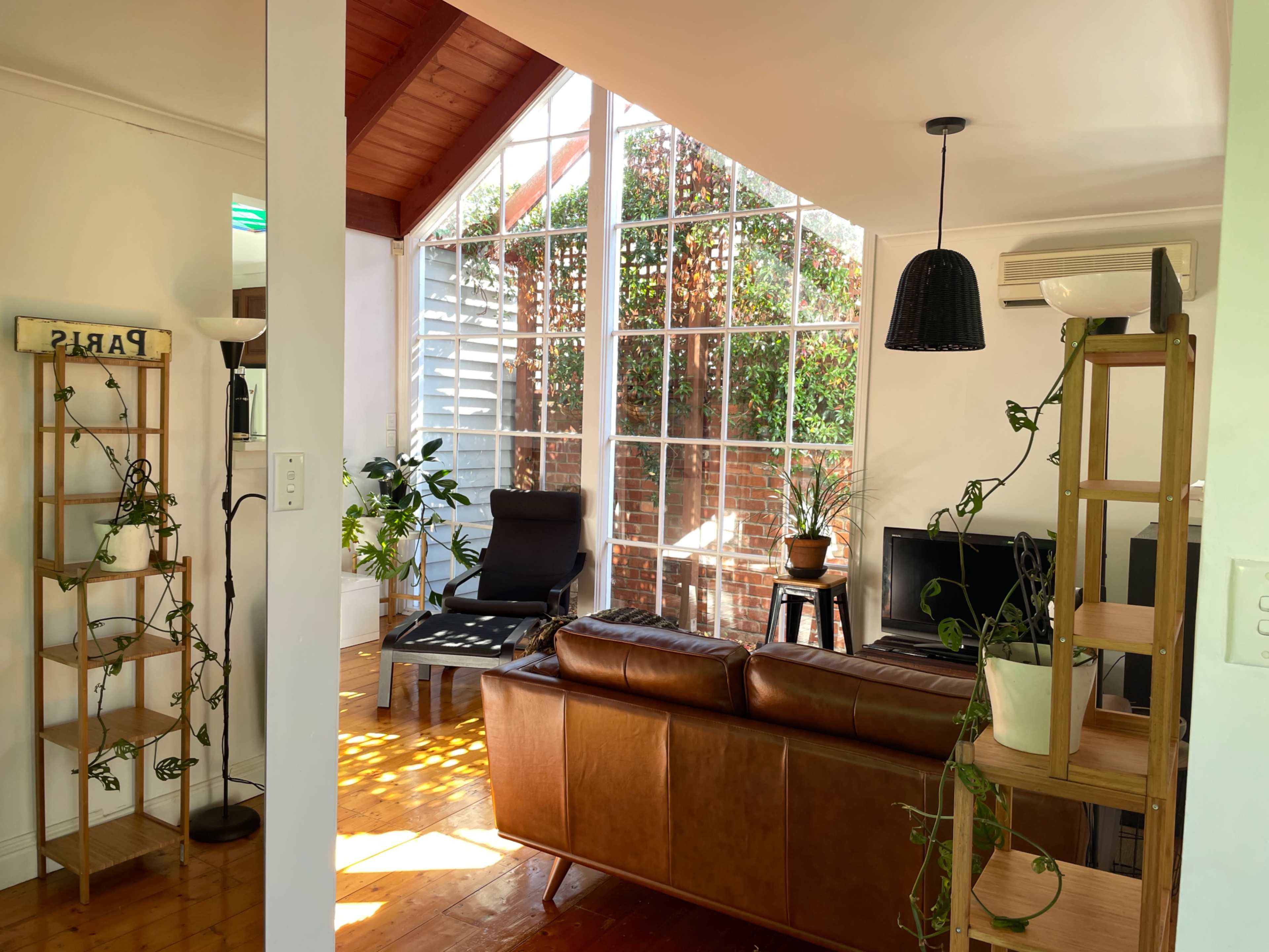 A bright living space with large glass windows, a brown leather sofa, a black chair, and various plants placed around the room.