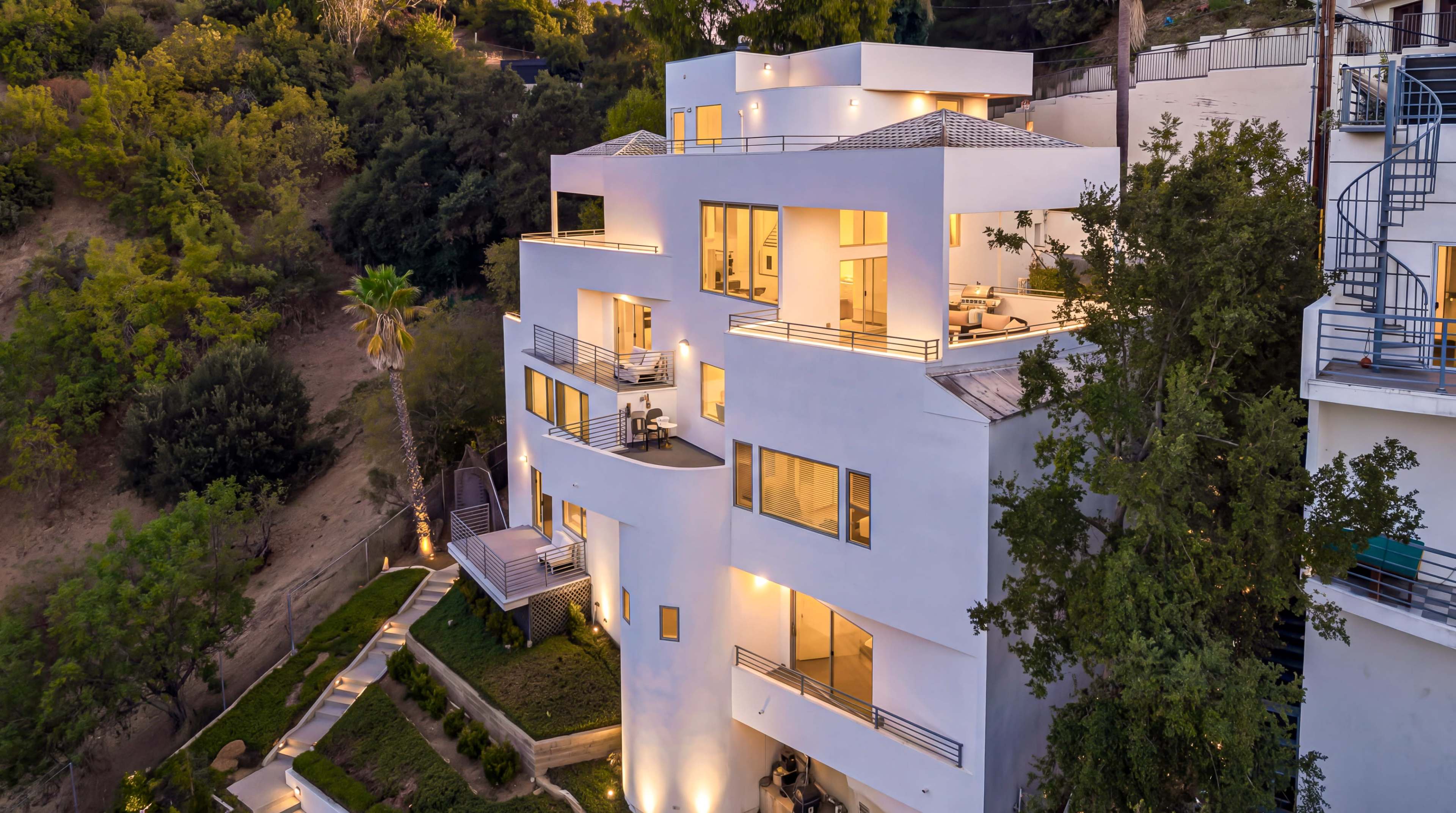 The image shows a modern white multi-level house with large windows, illuminated at dusk, situated on a hillside surrounded by greenery.