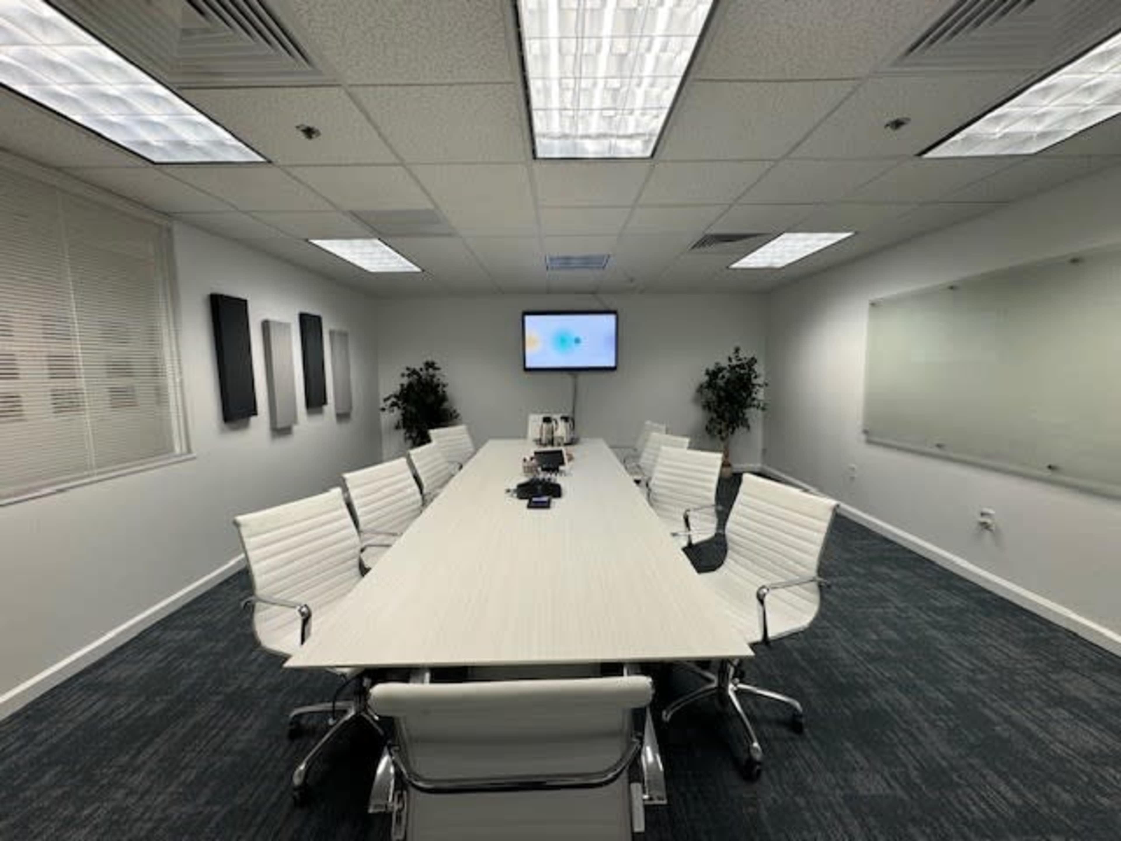 The image shows a modern conference room with a long white table, several white chairs, a wall-mounted screen, and potted plants.