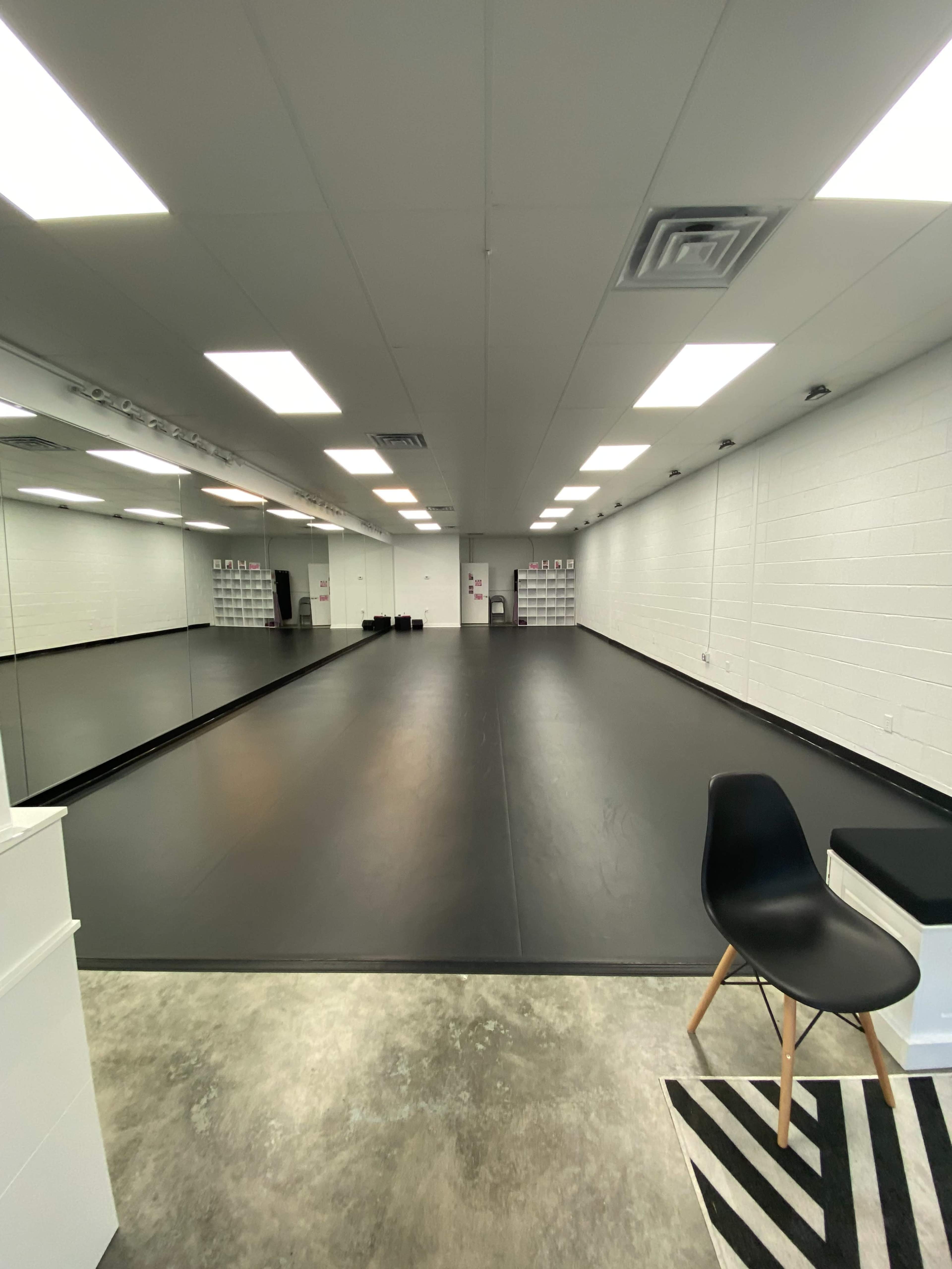 A large dance studio with a black floor, white walls, and bright overhead lighting.