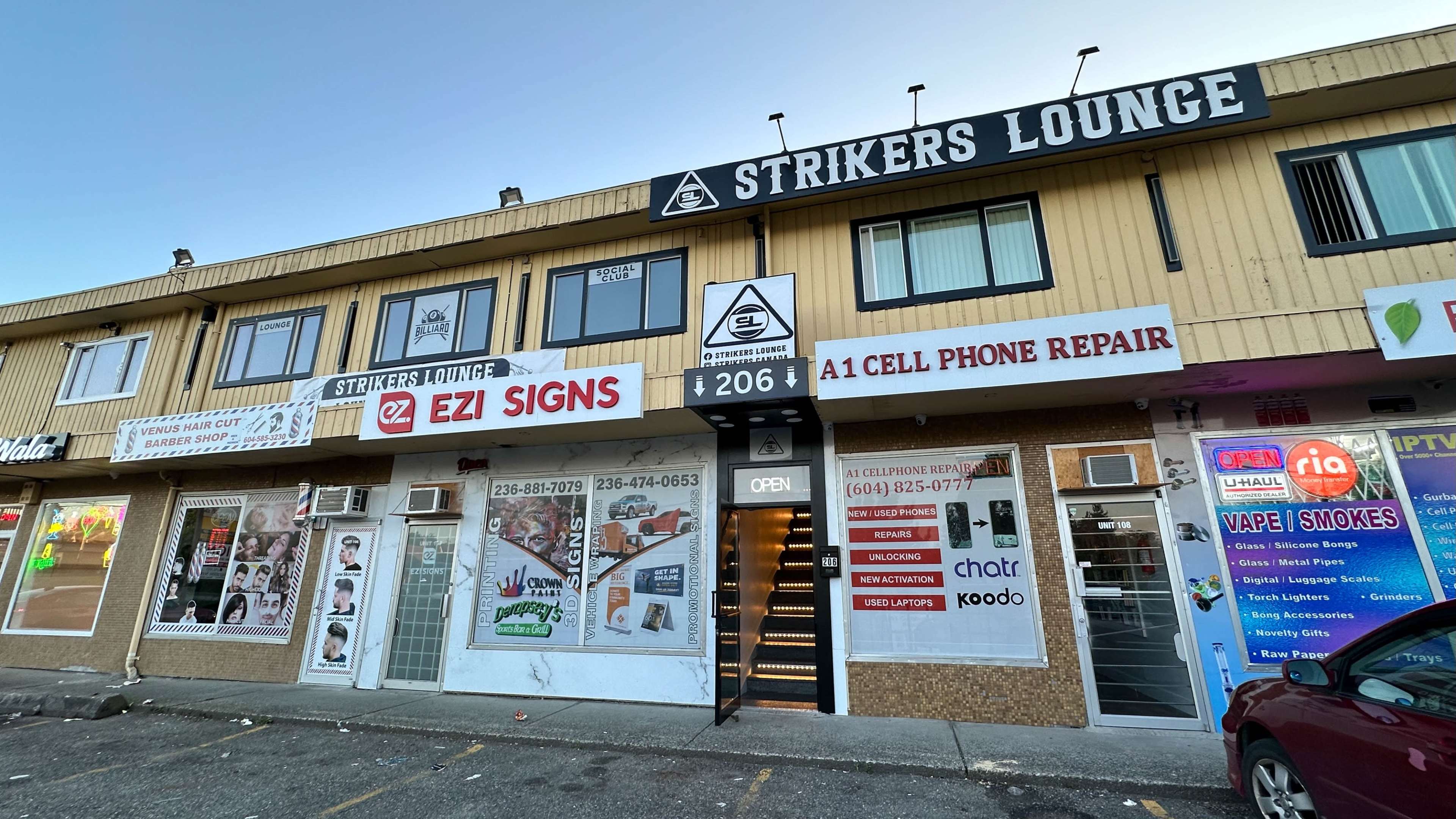 A row of businesses on a commercial building, featuring signs for Strikers Lounge, EZI Signs, and a cell phone repair shop, among others.