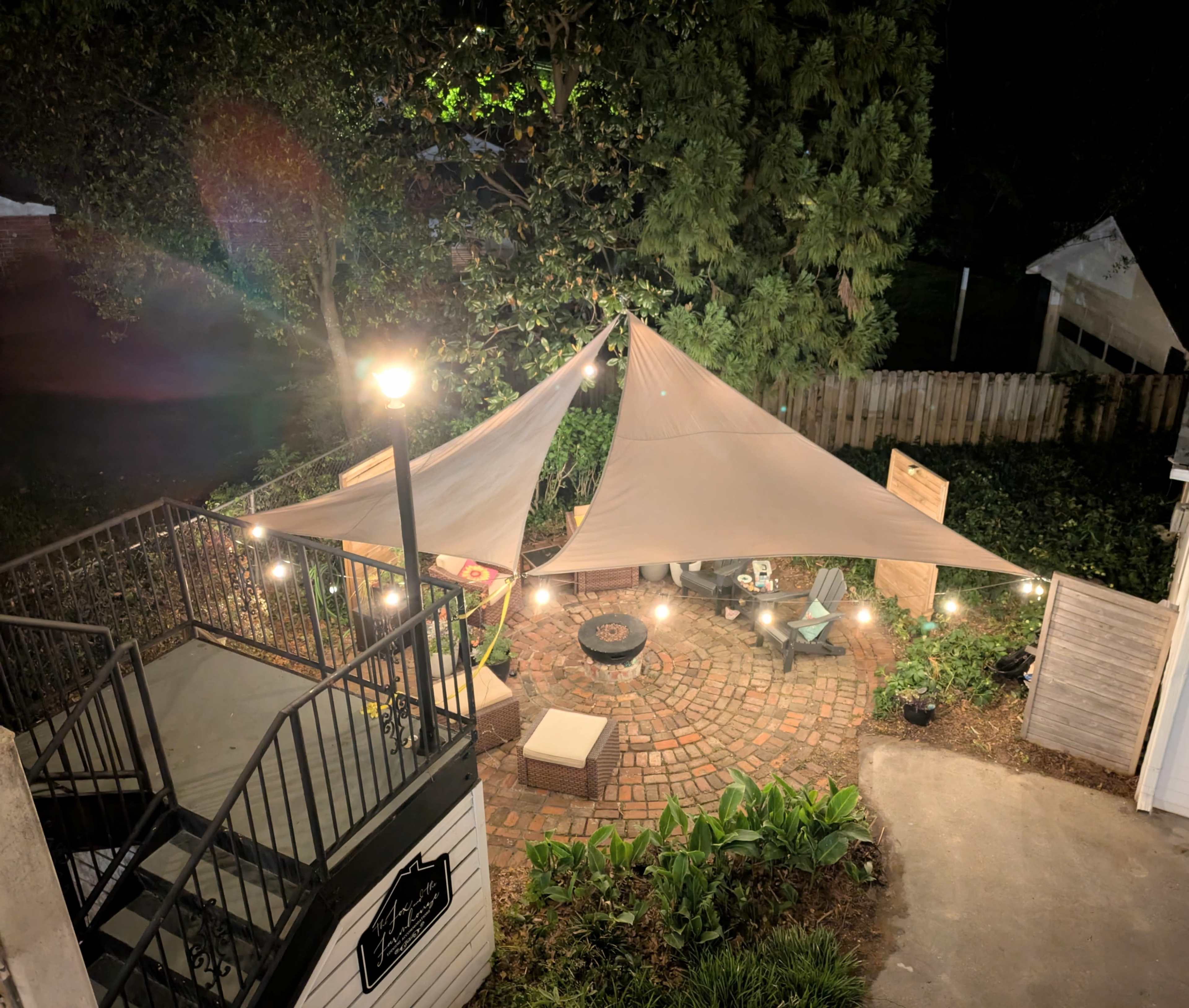 The image shows a backyard at night featuring a large, tan shade sail over a circular brick patio surrounded by outdoor furniture and string lights.
