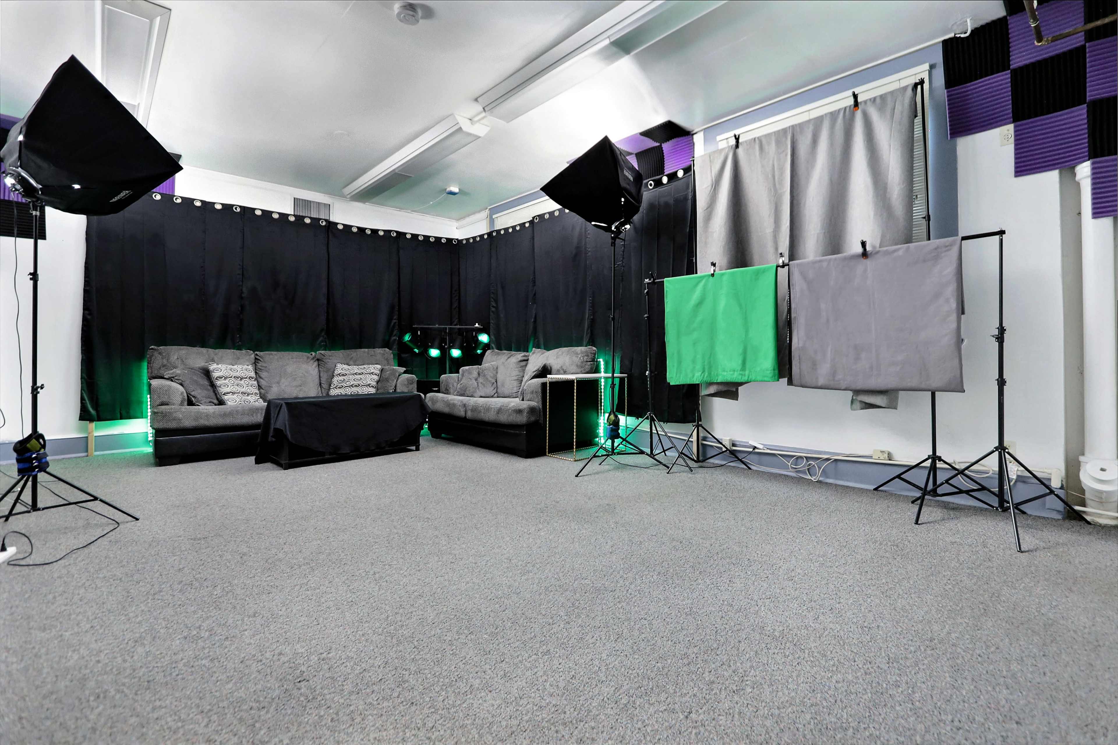A studio setup with a gray carpet, a black backdrop, a couch, and lighting equipment on both sides, along with green and gray backdrop panels on stands.
