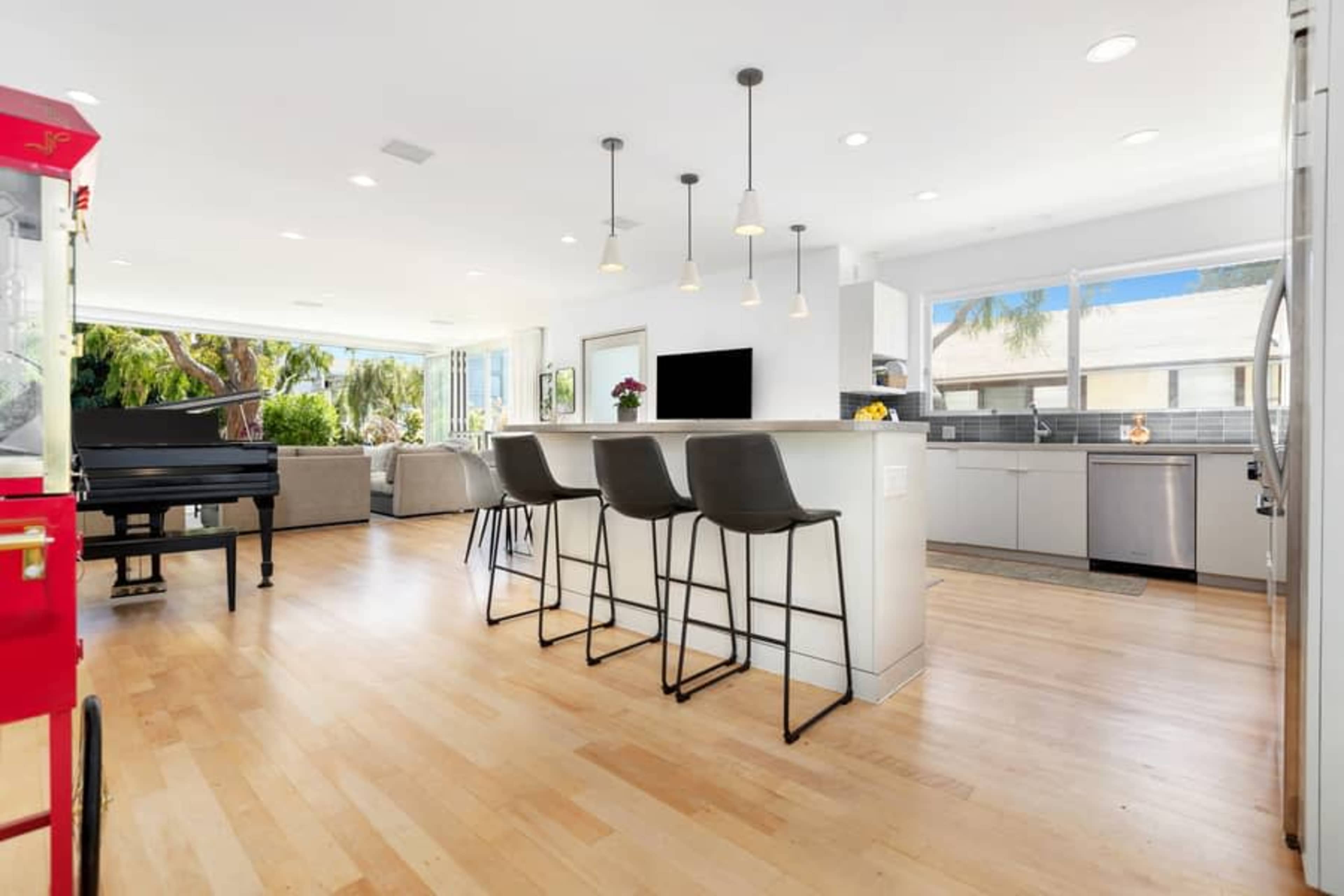 A modern kitchen and living area with a central island and seating, large windows, and a grand piano in the corner.