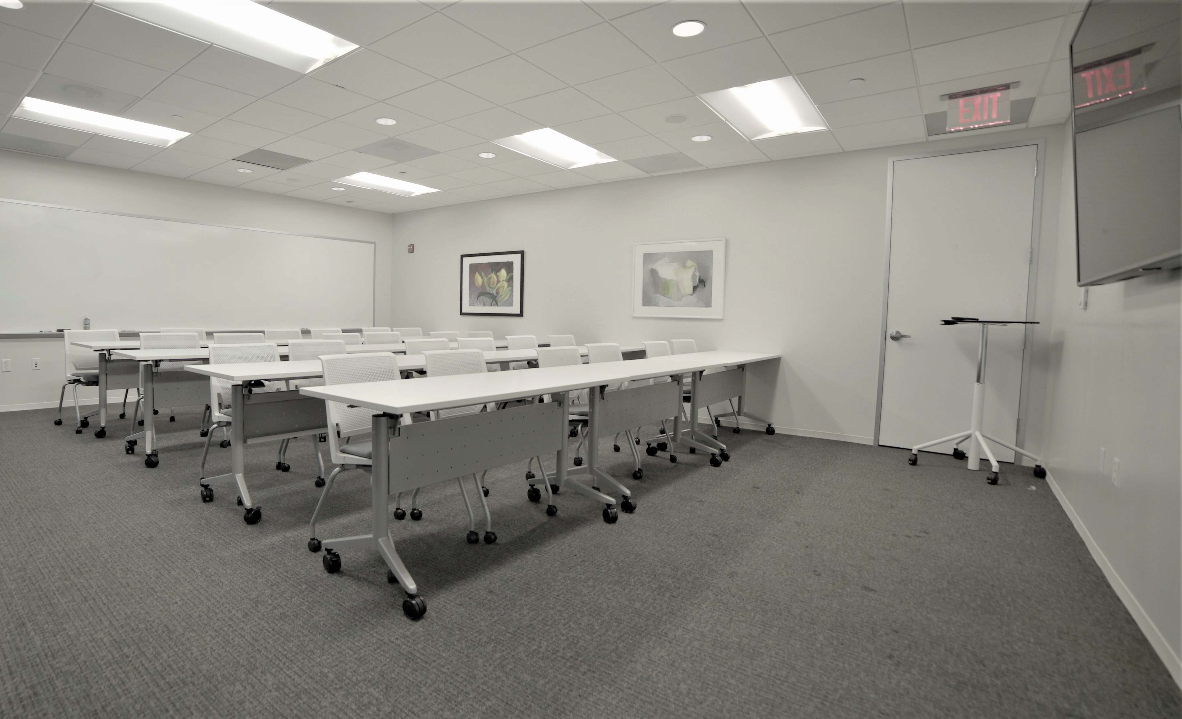 A modern, empty classroom arranged with rows of movable tables and chairs, featuring white walls and overhead lighting.