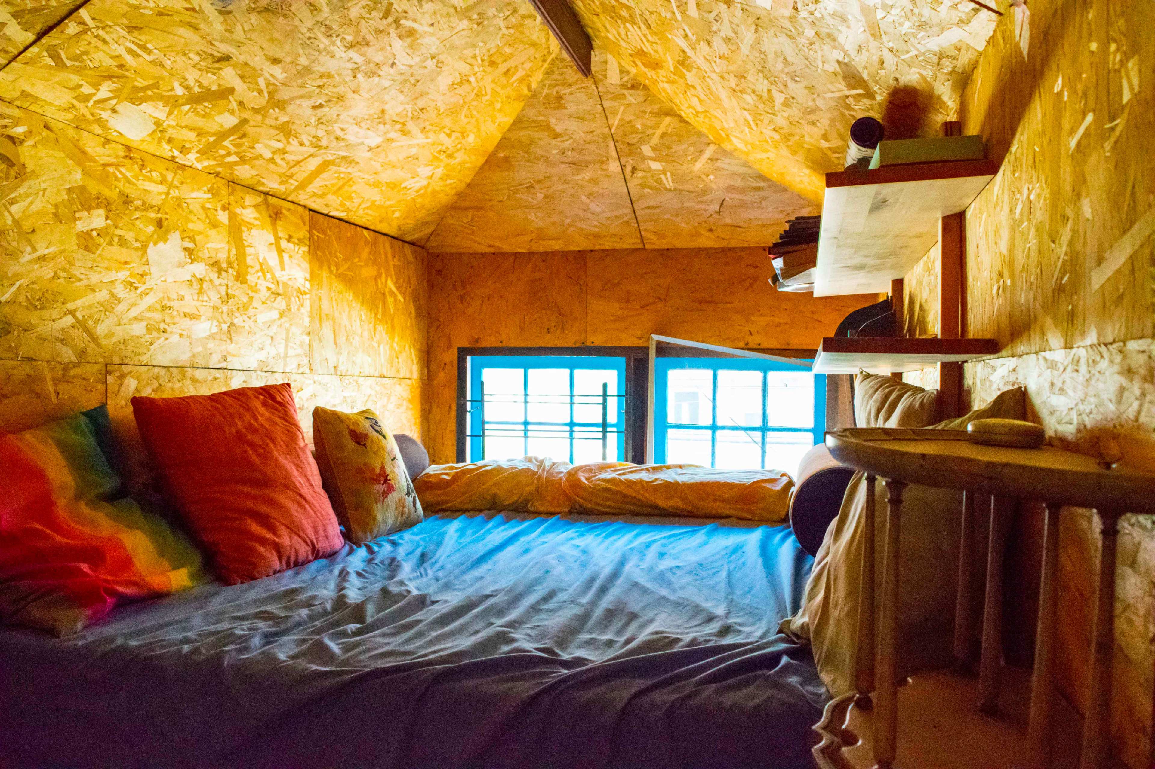 A cozy loft space with a bed, colorful pillows, and wooden shelves, featuring large windows that let in natural light.