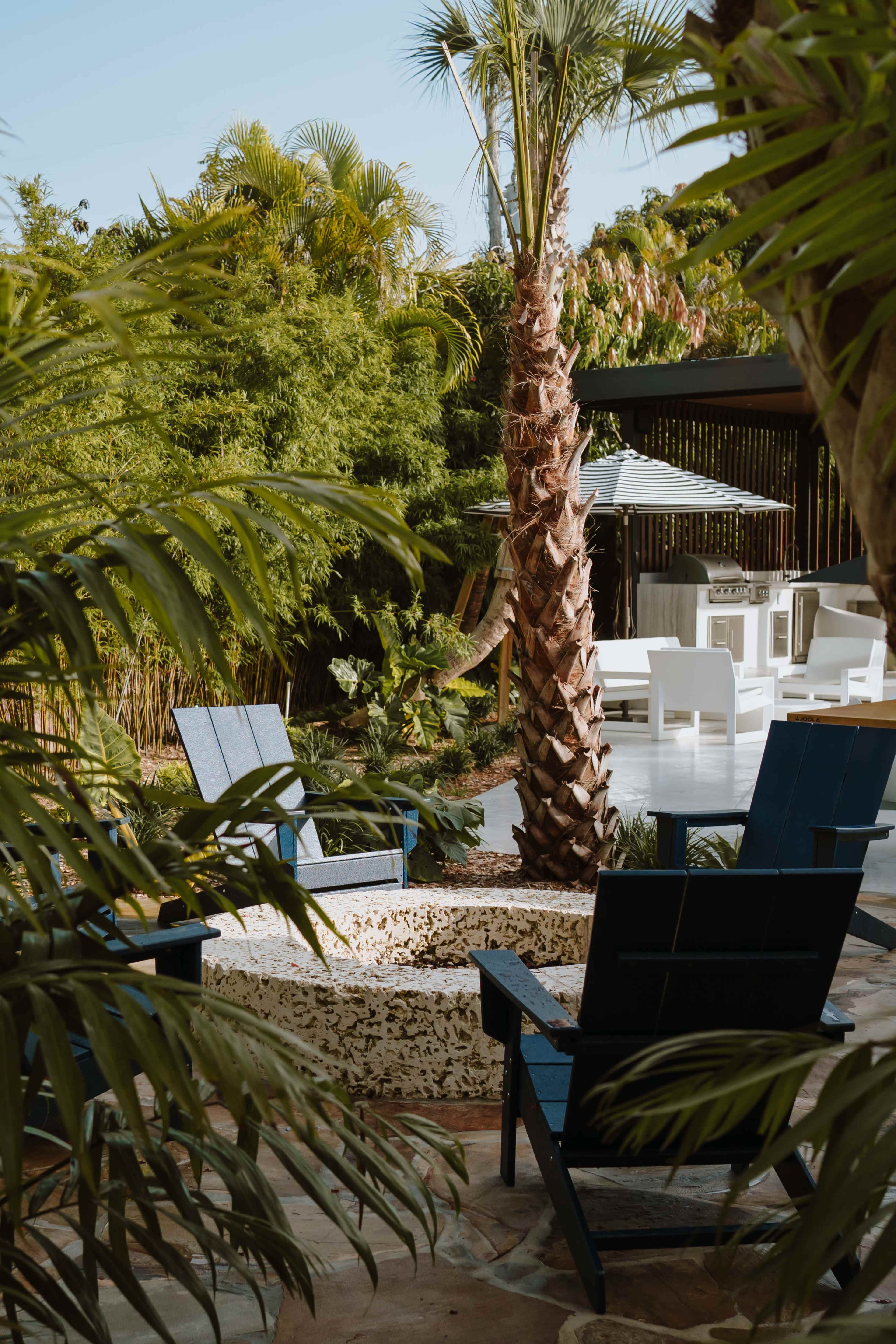 The image shows a serene outdoor space featuring a fire pit surrounded by two blue chairs, with lush tropical plants and a palm tree in the background.