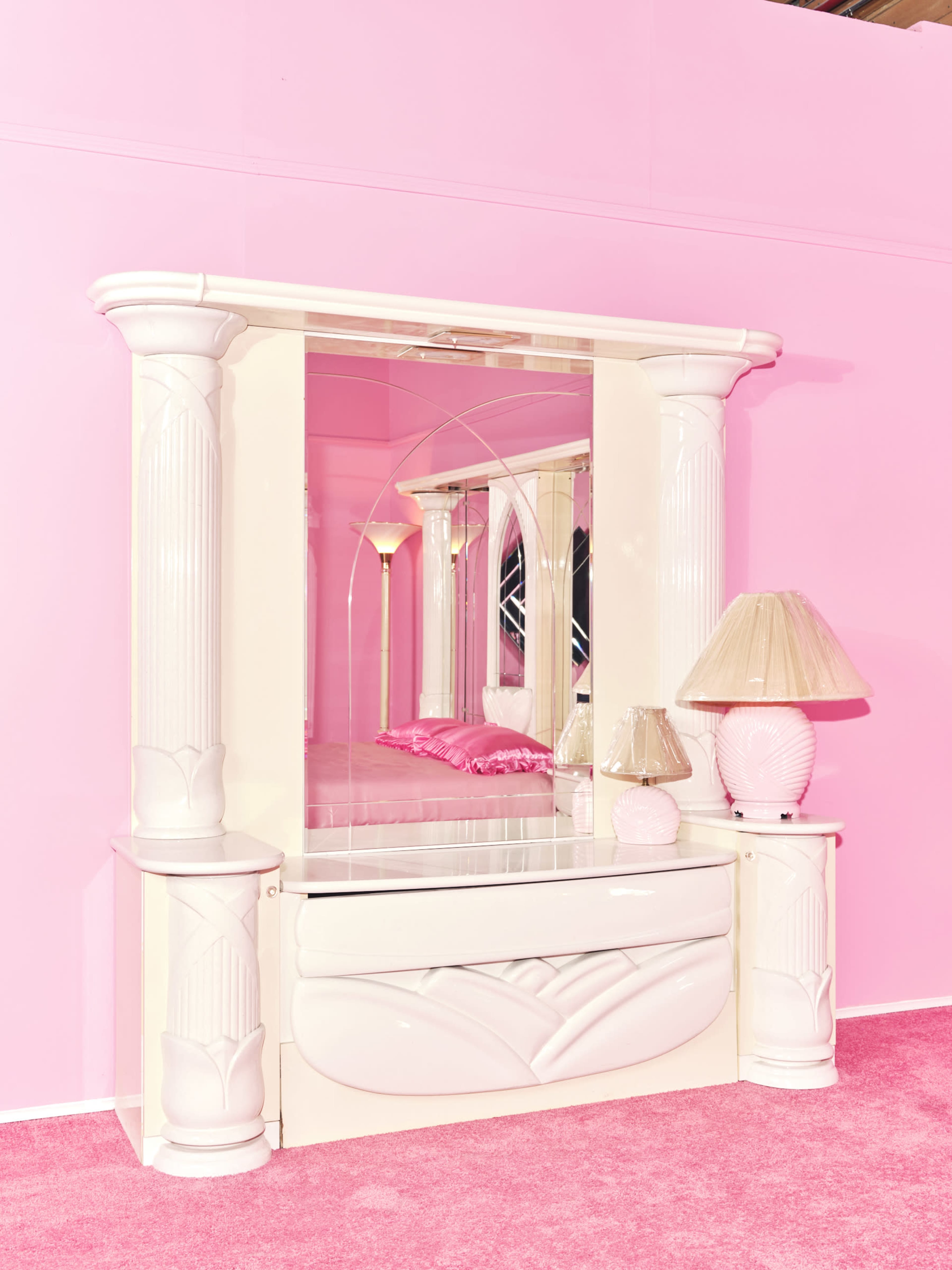 The image depicts a white dresser with a large mirror and decorative columns, set against a pink background and accompanied by a pink lamp.