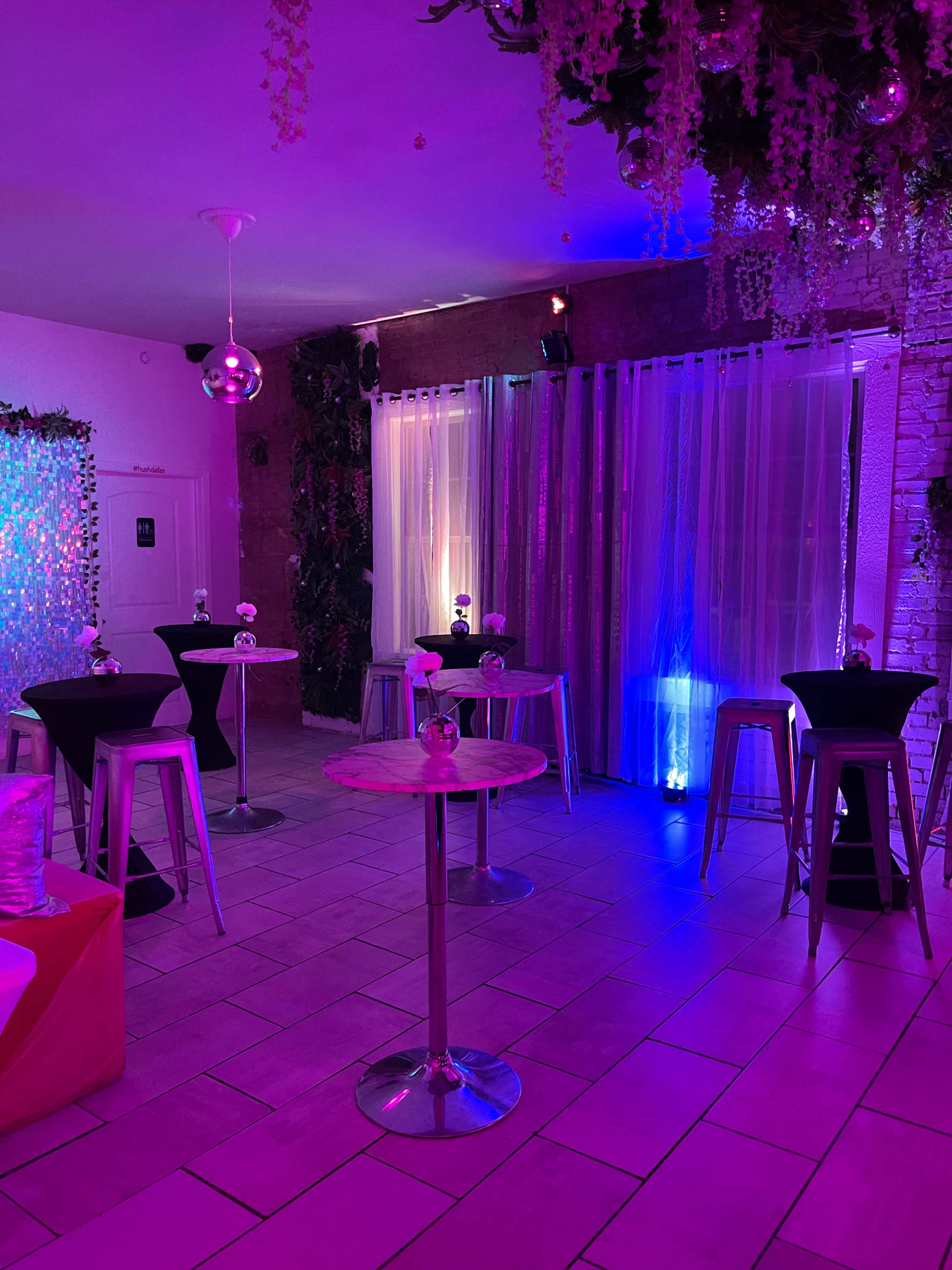 A dimly lit event space features tables with black and silver coverings, illuminated by purple and blue lights, alongside decorative plants and sheer curtains.