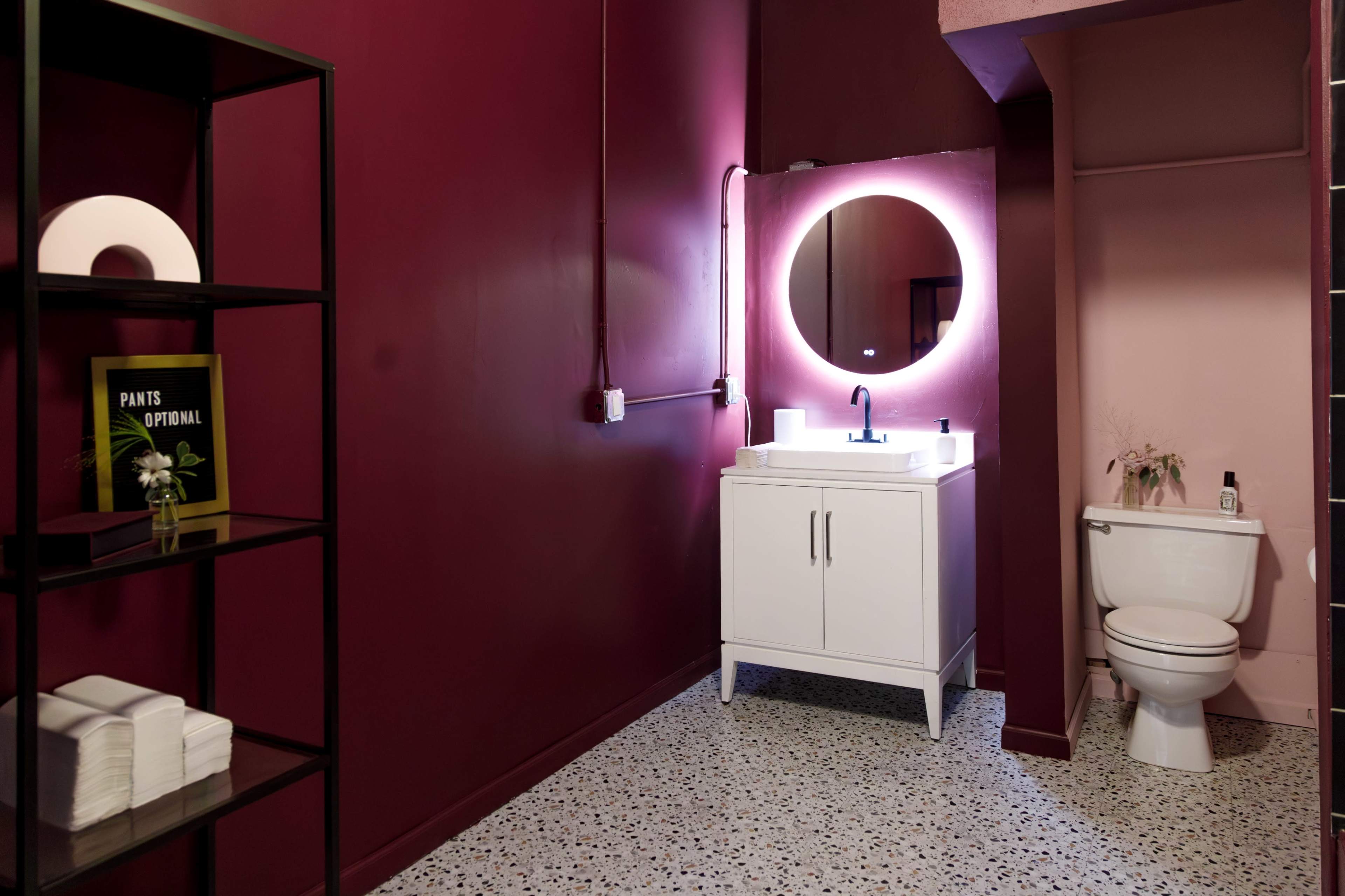 A modern bathroom with a white vanity, mirror, toilet, and a black shelf displaying folded towels and a book against a maroon wall.