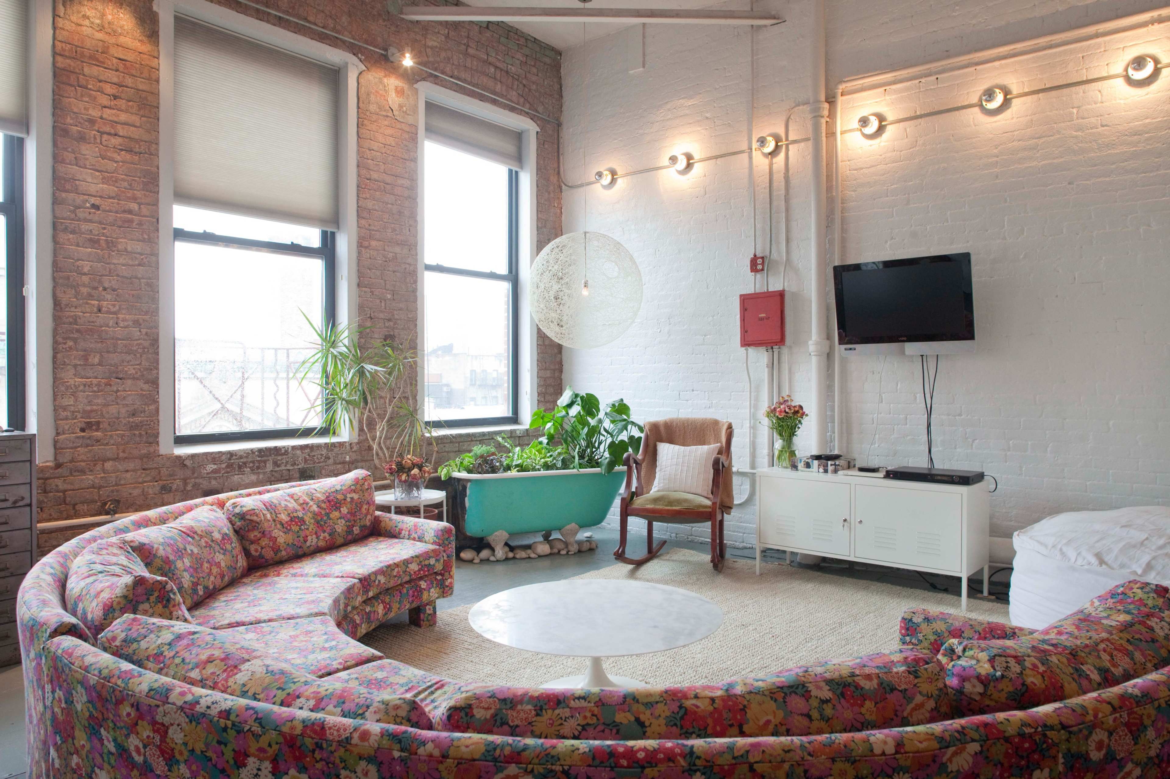 A living room features a curved floral-patterned sofa, a round table, a hanging light fixture, and large windows with plants in a blue tub.