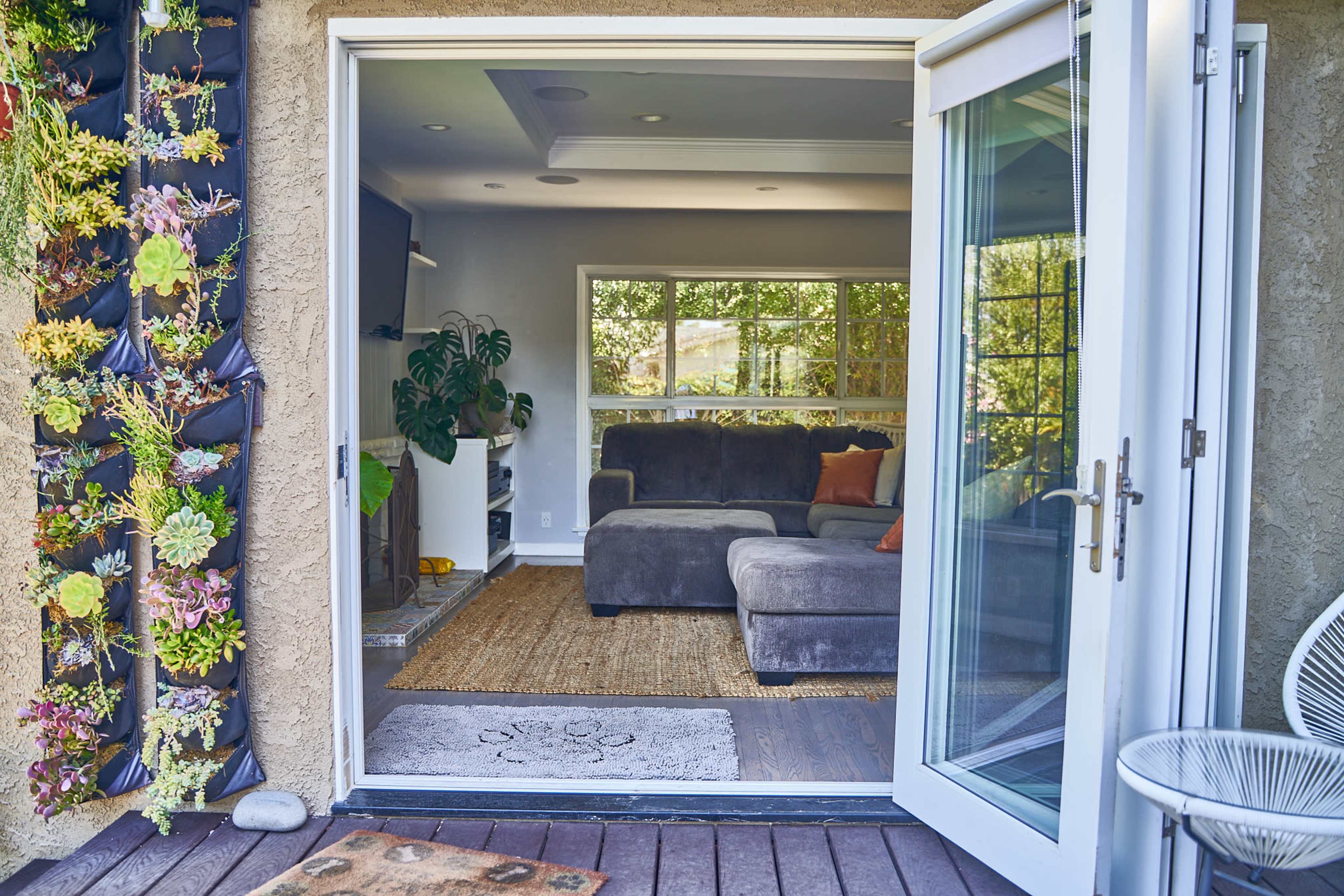 The image shows an open doorway leading from a deck into a living room with a gray sectional sofa, a large plant, and a decorative wall of succulents.
