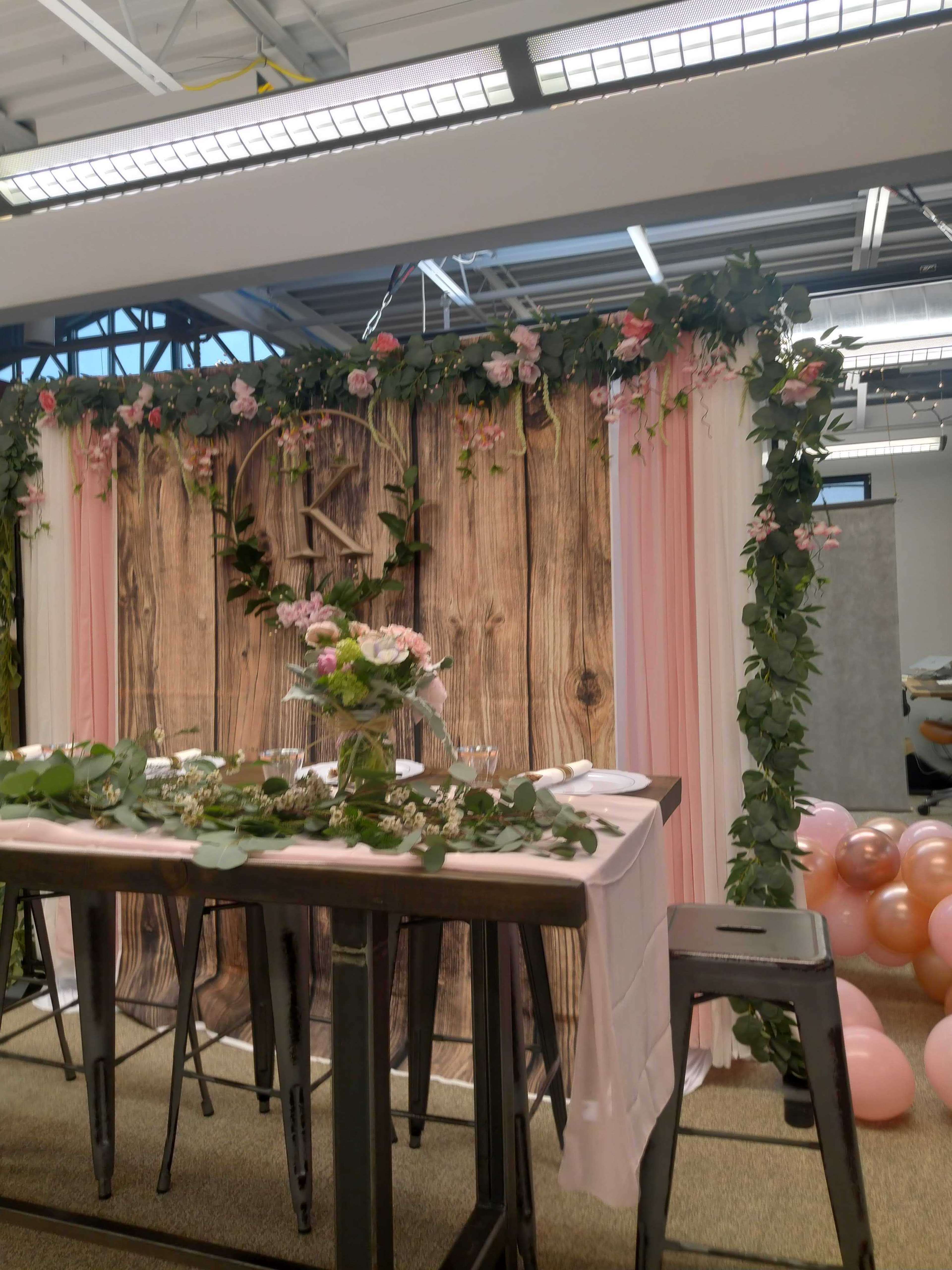 The image shows a decorated table set for an event, featuring a wooden backdrop adorned with flowers and greenery, surrounded by pastel balloons.