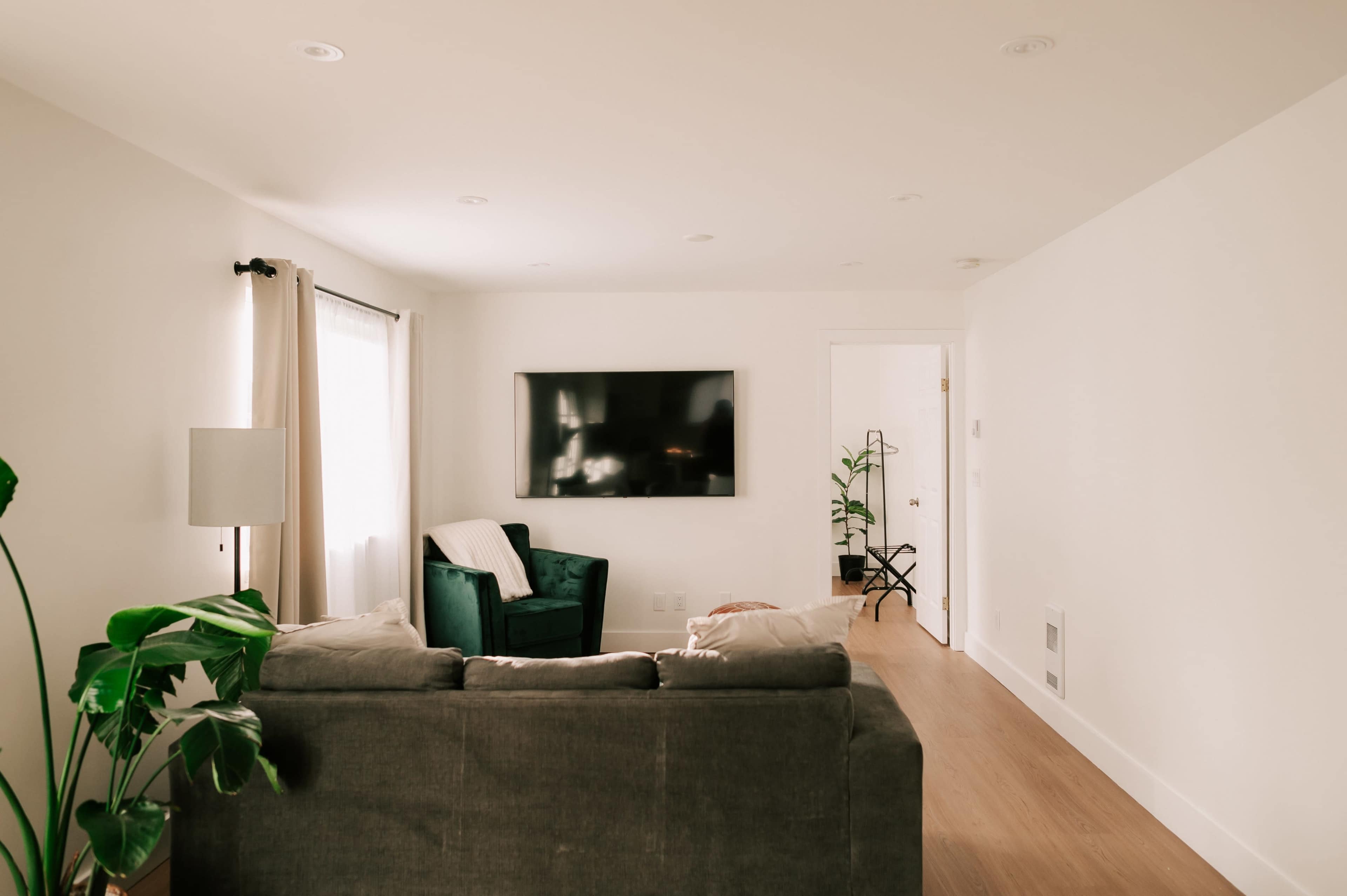 The image shows a modern living room with a gray couch facing a large wall-mounted television and a green armchair beside a doorway.