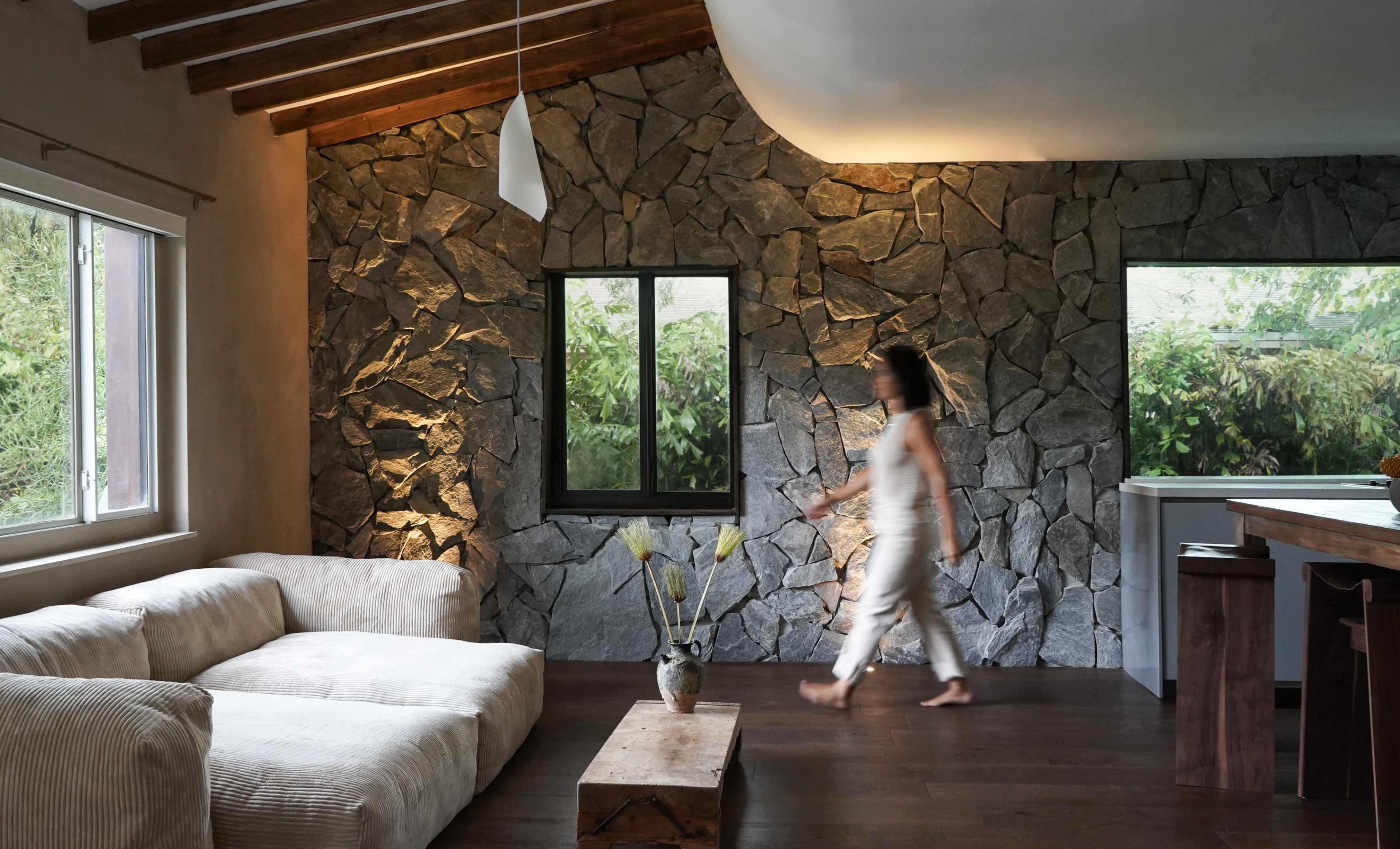A woman walks across a living room featuring a stone wall, large windows, and a light-colored sofa.