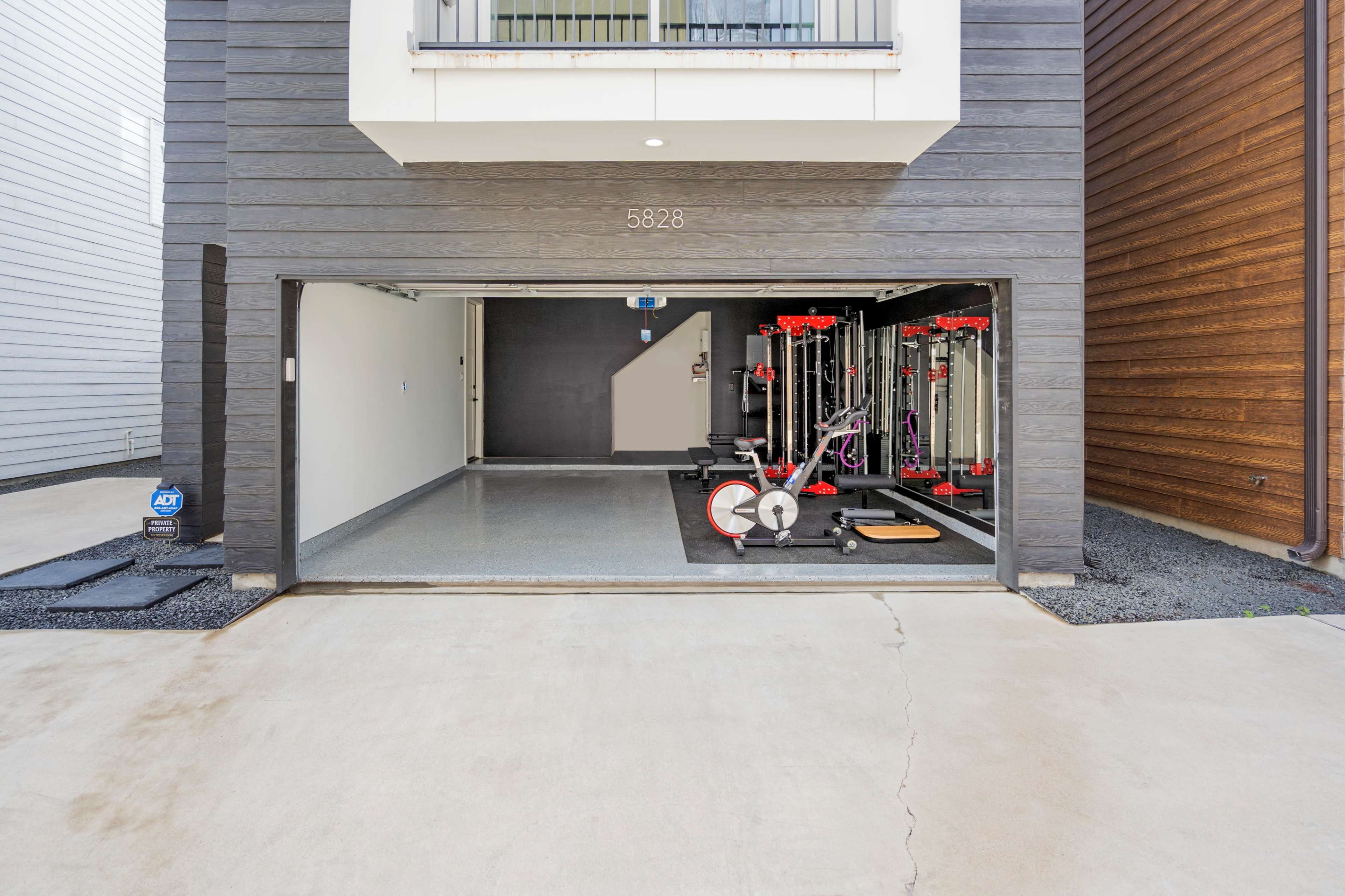 An open garage with workout equipment neatly arranged inside, featuring various machines and weights against a gray wall.