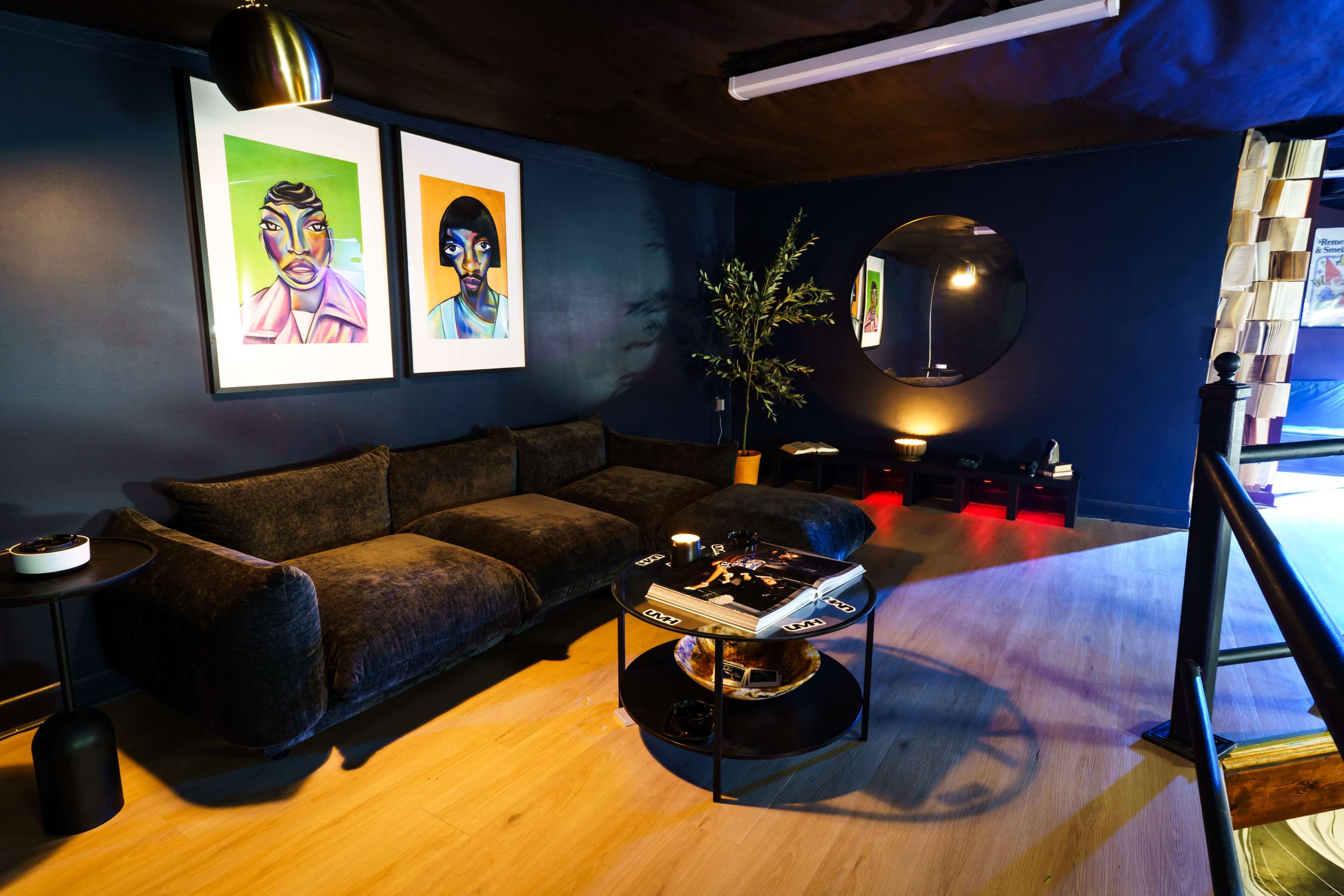 The image shows a modern lounge area with dark blue walls, a plush sofa, and a round mirror, adorned with vibrant artwork and decorative elements.