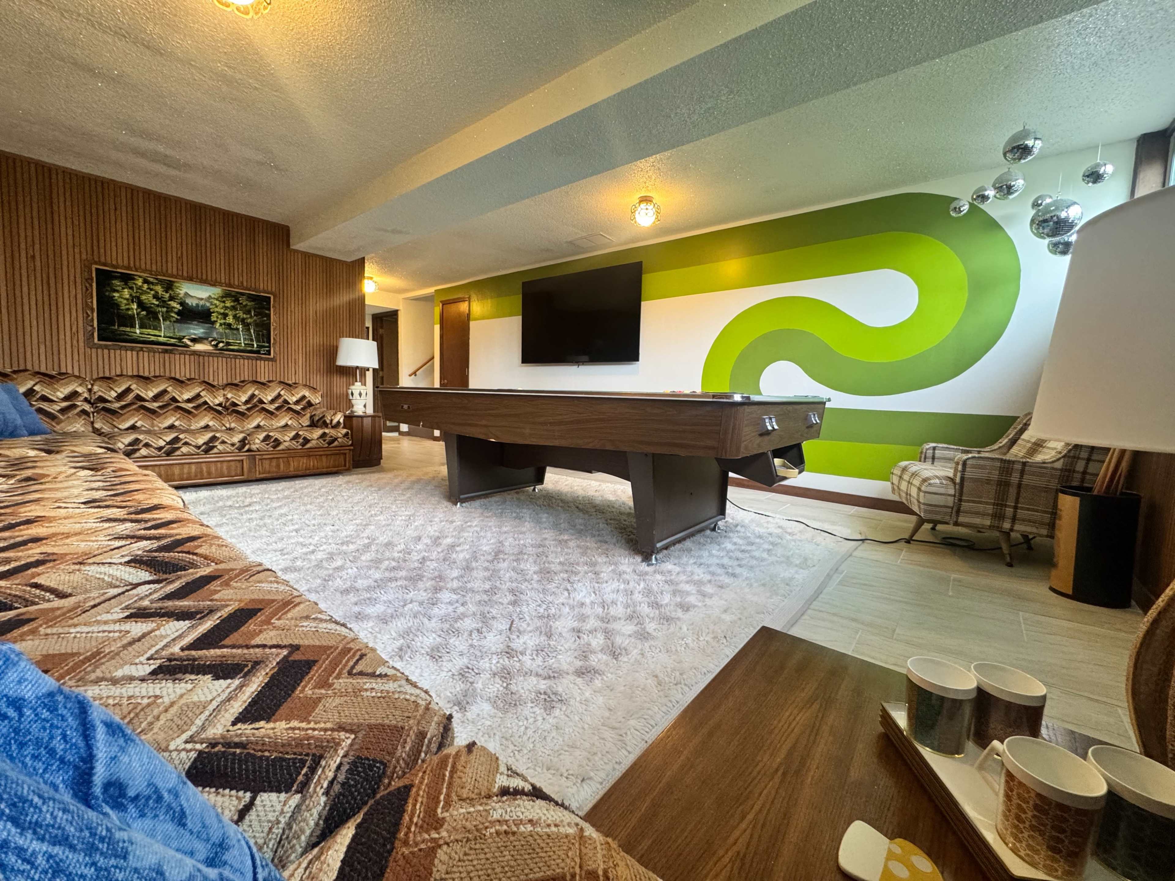 The image shows a modern game room featuring a pool table at the center, with a green abstract mural on the wall and retro-style furniture around the space.