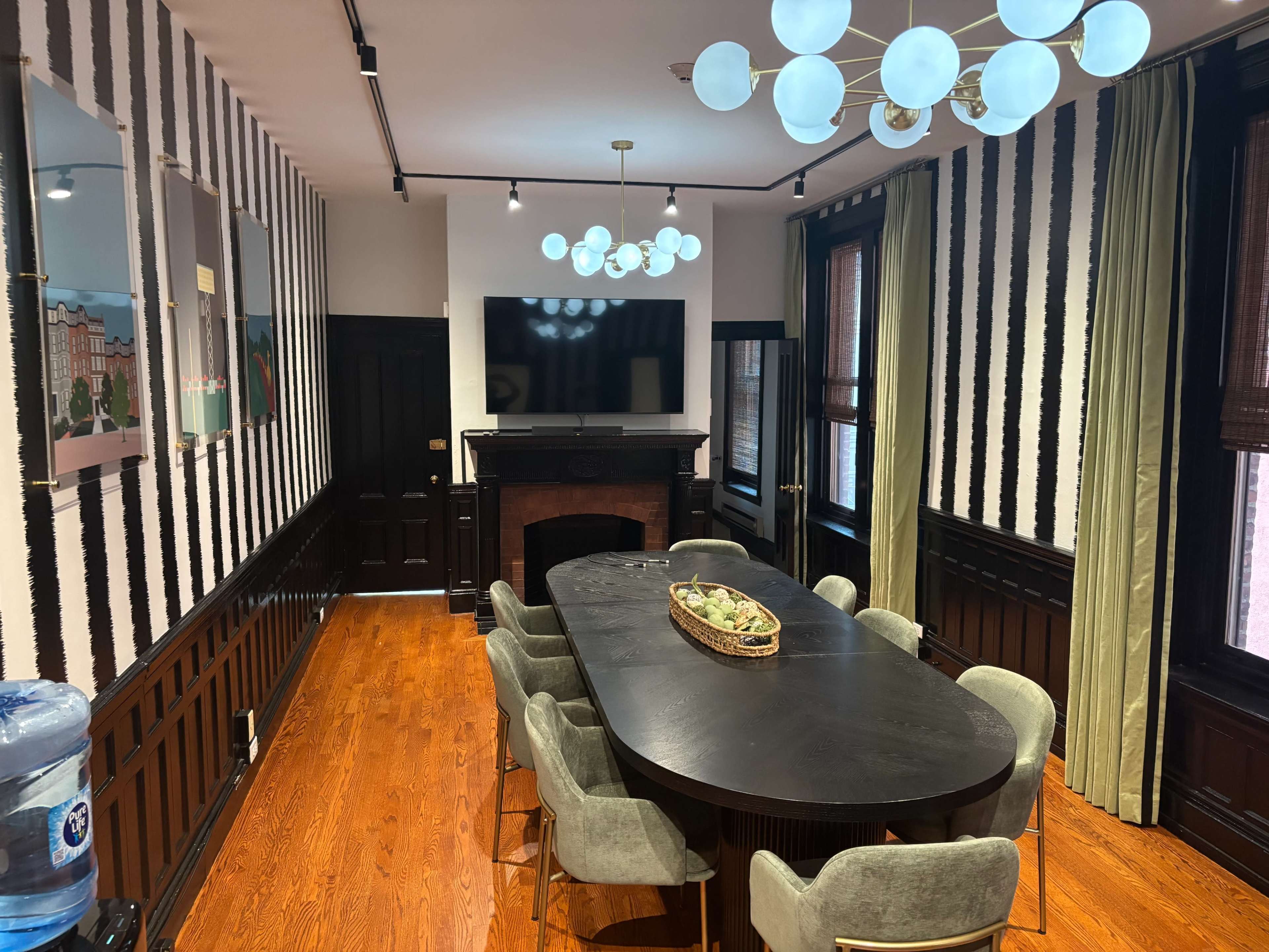 The image shows a modern dining room featuring a long black table surrounded by green upholstered chairs, with a stylish light fixture overhead and a television mounted on the wall.