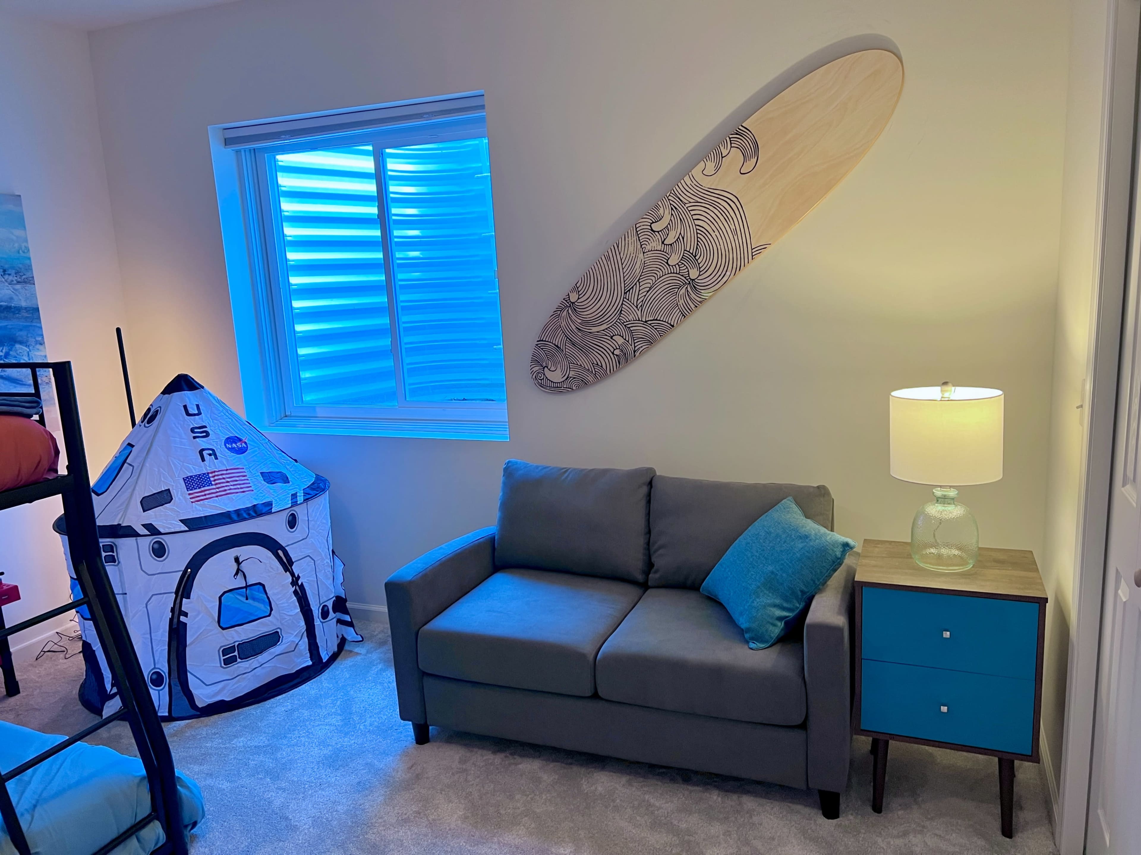 The image shows a cozy room featuring a gray sofa, a blue side table with a lamp, a playful tent shaped like a space capsule, and a decorative surfboard on the wall near a window.