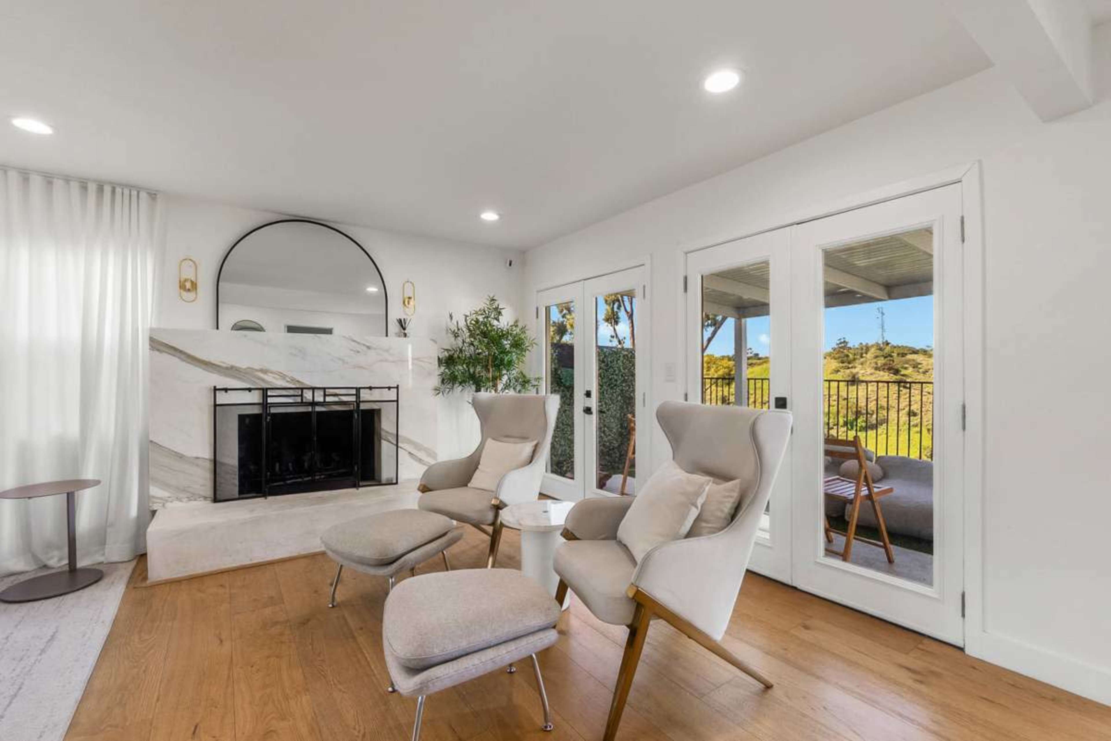 The image shows a modern living room featuring two stylish armchairs positioned next to a small round table, a fireplace with a marble facade, and sliding glass doors that lead to an outdoor space.