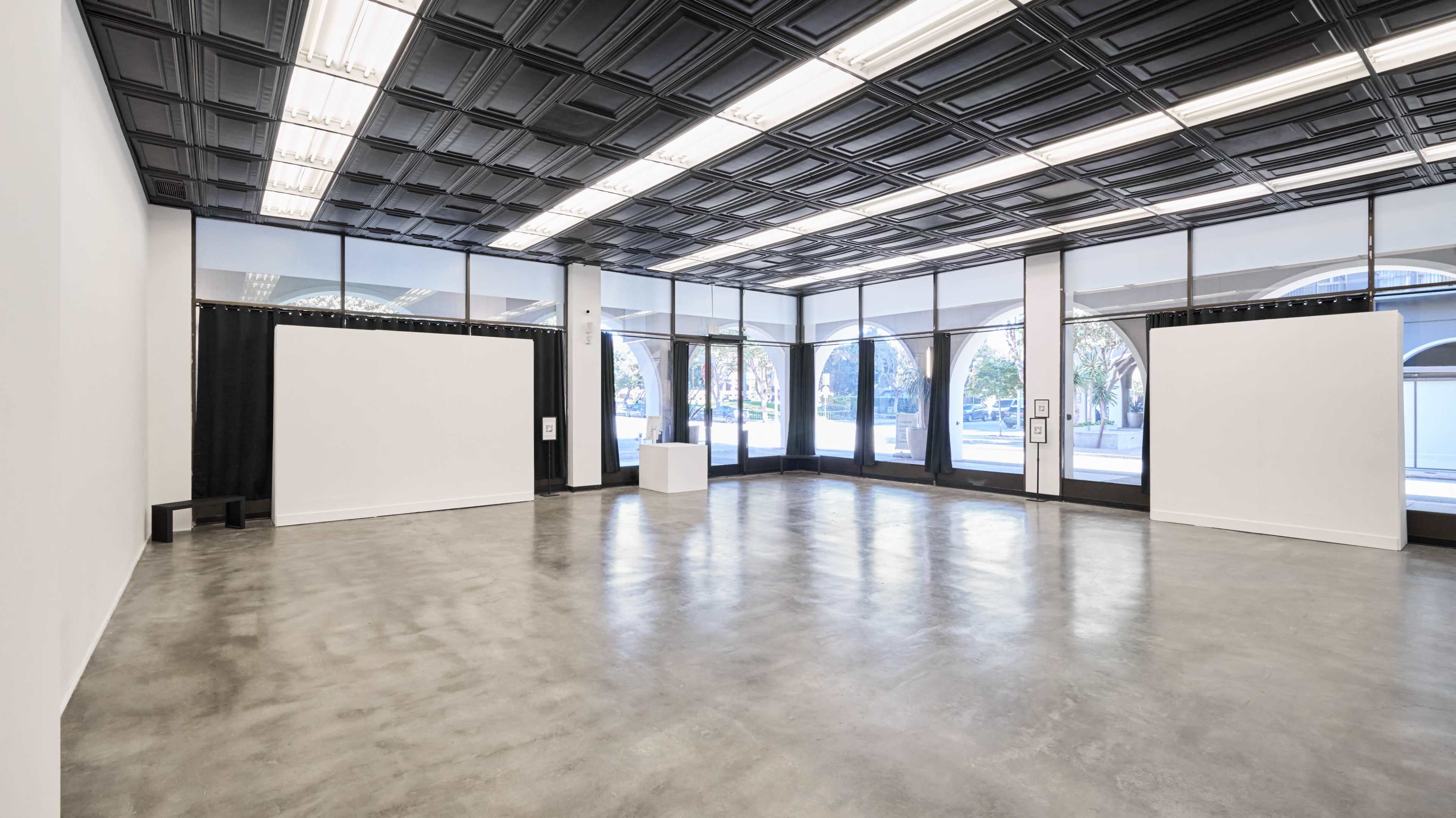 The image shows an empty art gallery with large windows, white walls, and concrete flooring.