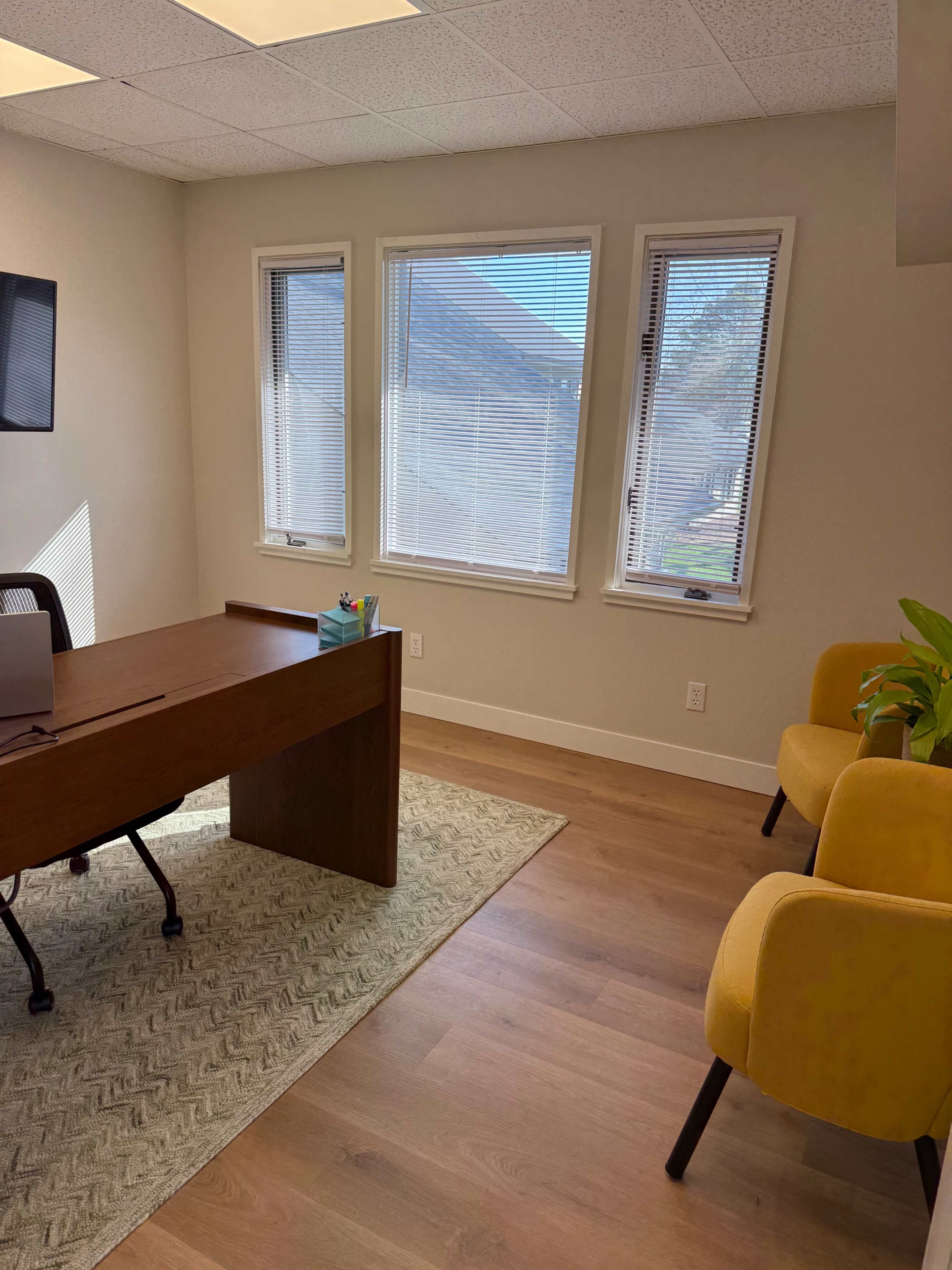 The image shows a bright office space with a wooden desk, two yellow chairs, and large windows allowing natural light to enter.