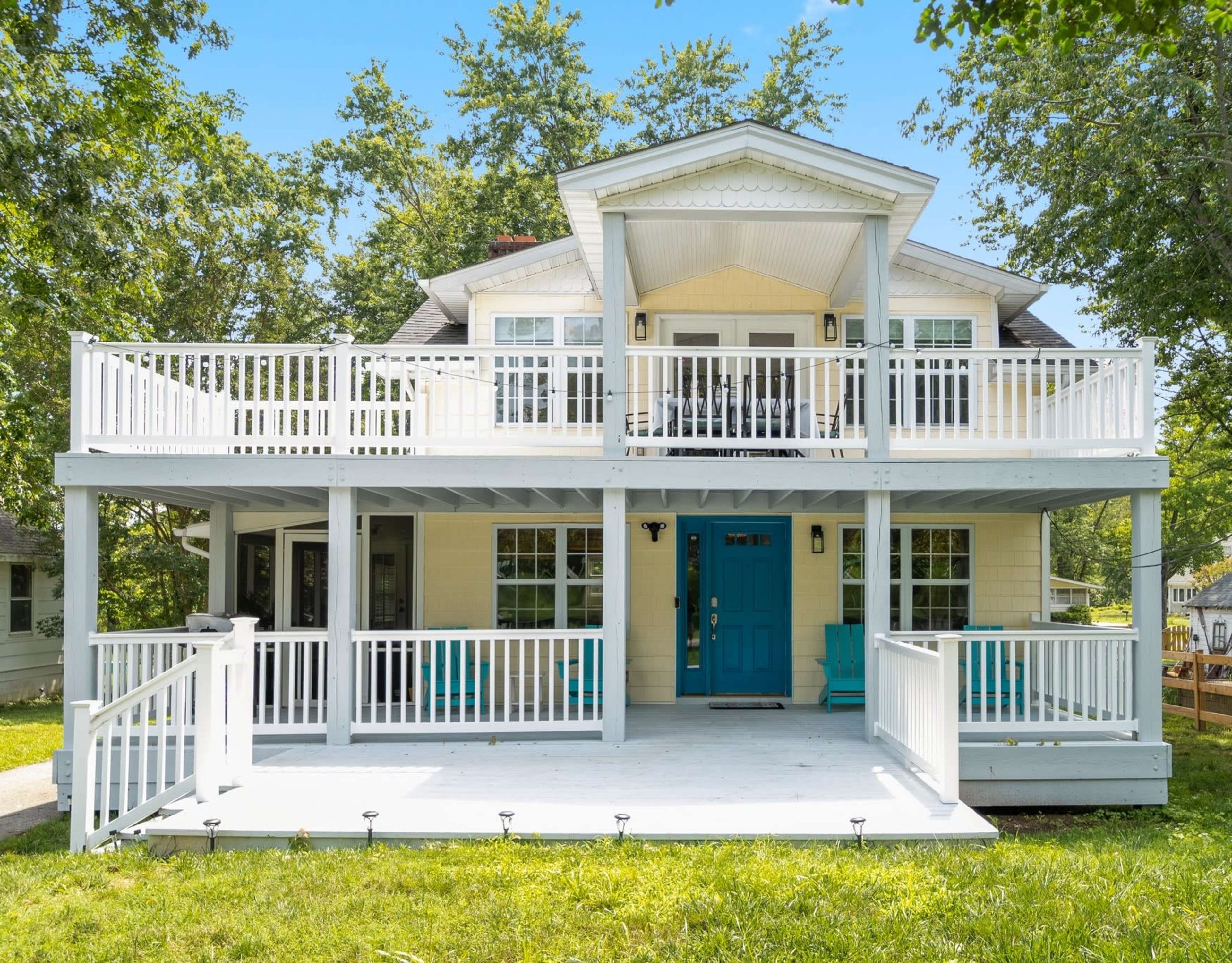 A two-story house with a wraparound porch and a blue front door is surrounded by greenery.