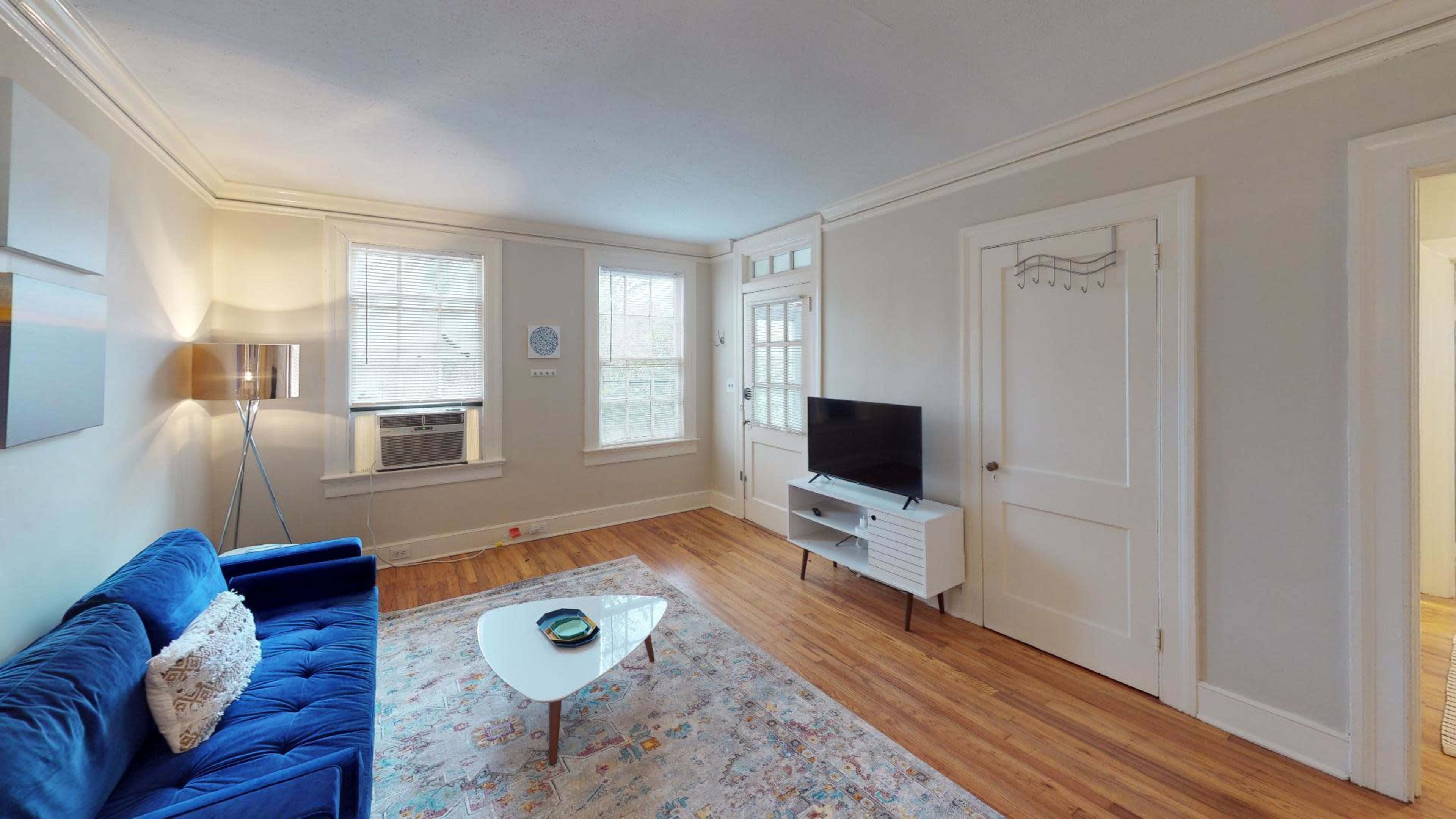 The image shows a living room with a blue couch, a coffee table, a television on a white stand, and large windows that allow natural light to fill the space.