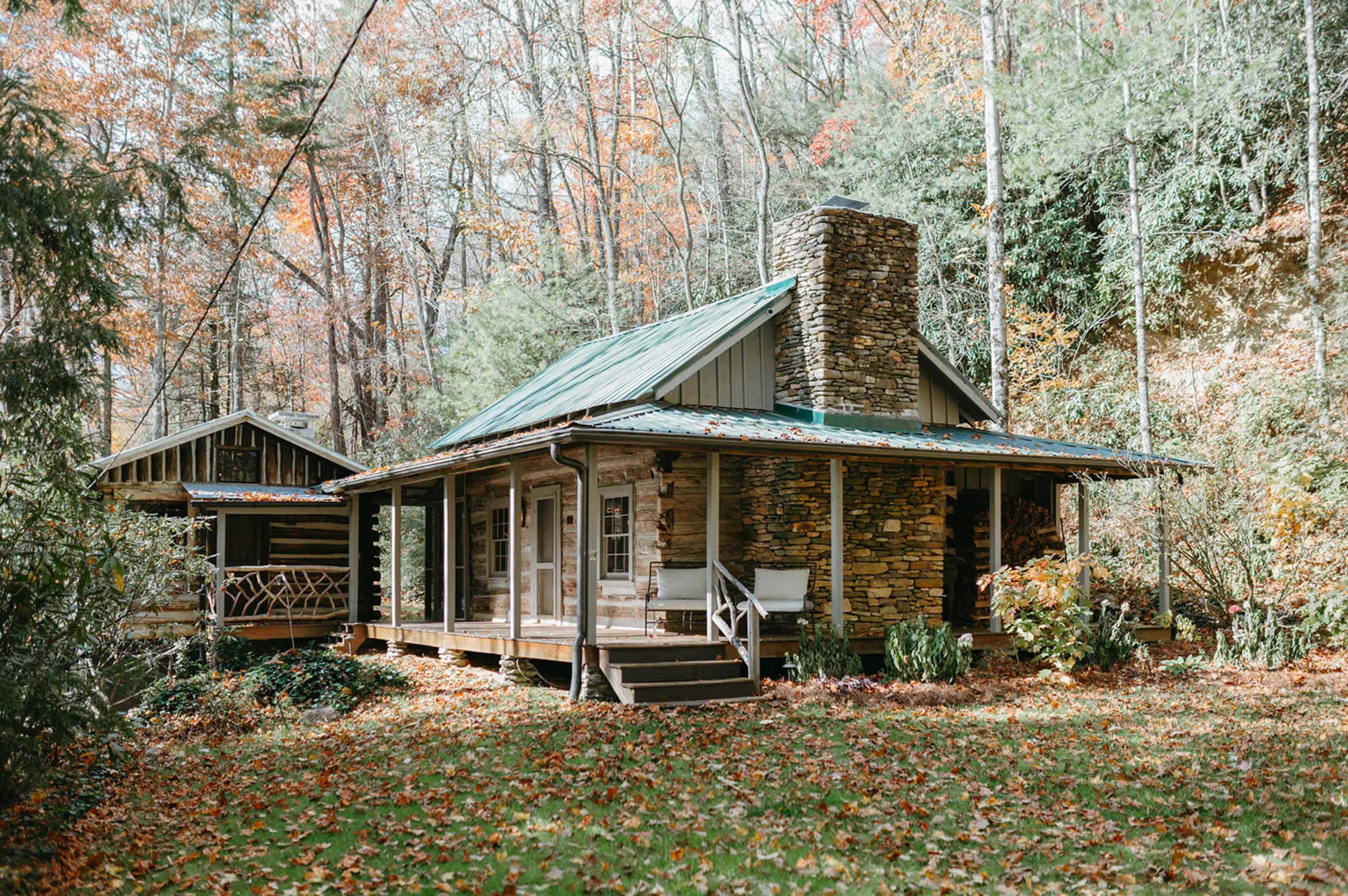 A stone cottage with a metal roof is surrounded by colorful autumn foliage and features a chimney and a nearby log cabin.