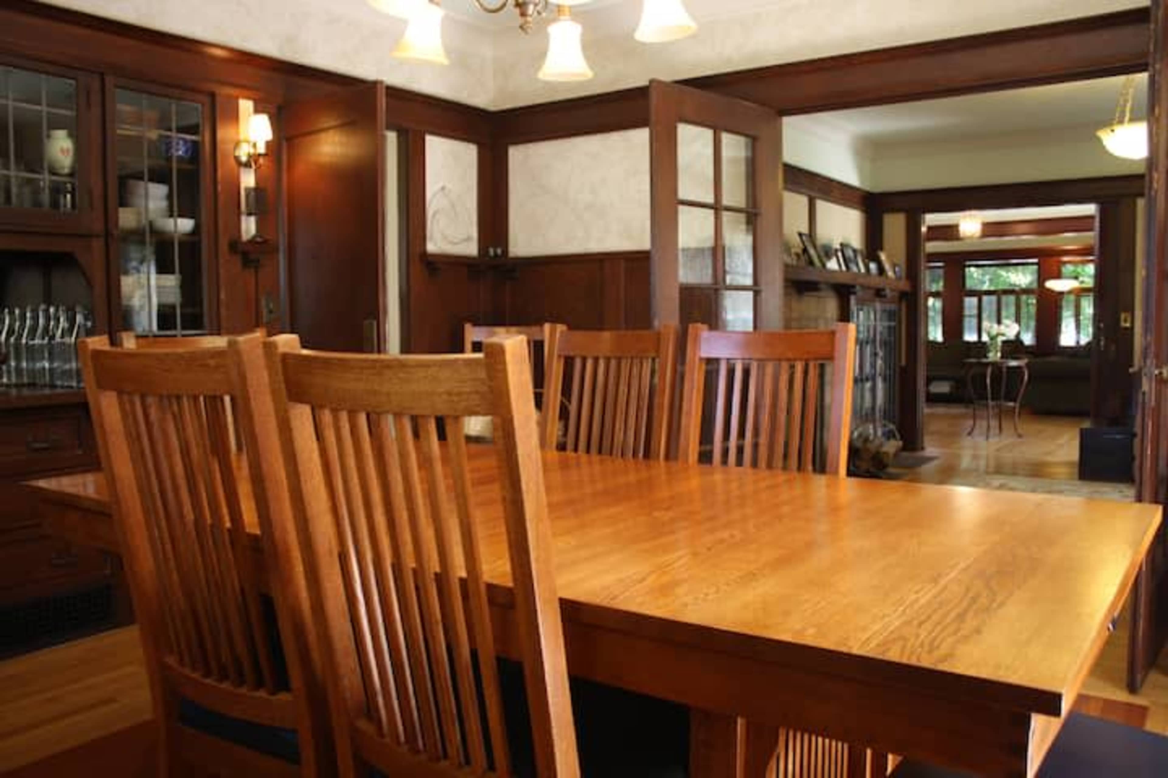 A wooden dining table is set with several upright wooden chairs in a well-lit room featuring a built-in cabinet and large windows.