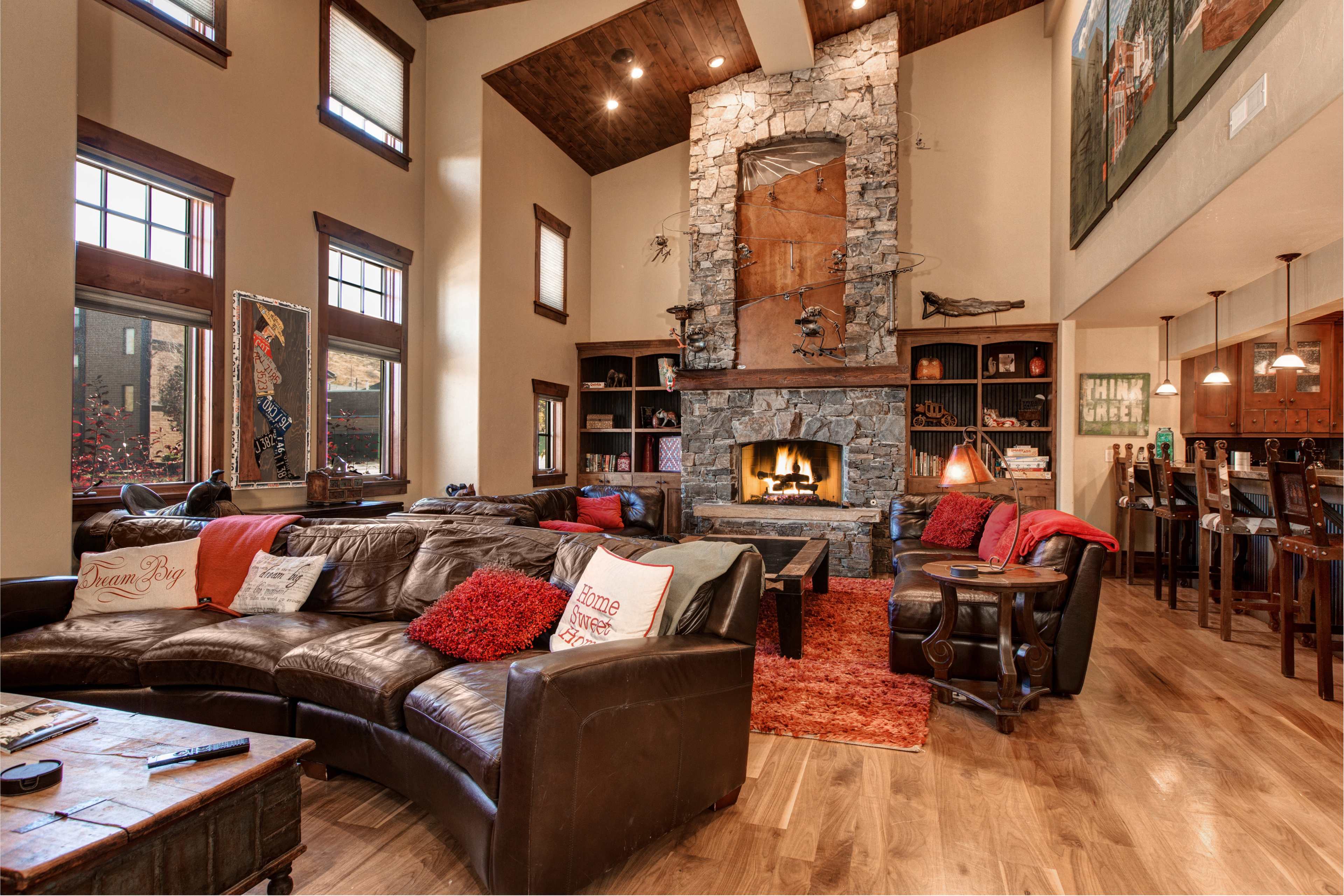 A spacious living room with a stone fireplace surrounded by a leather sectional sofa, wooden ceiling beams, and large windows.