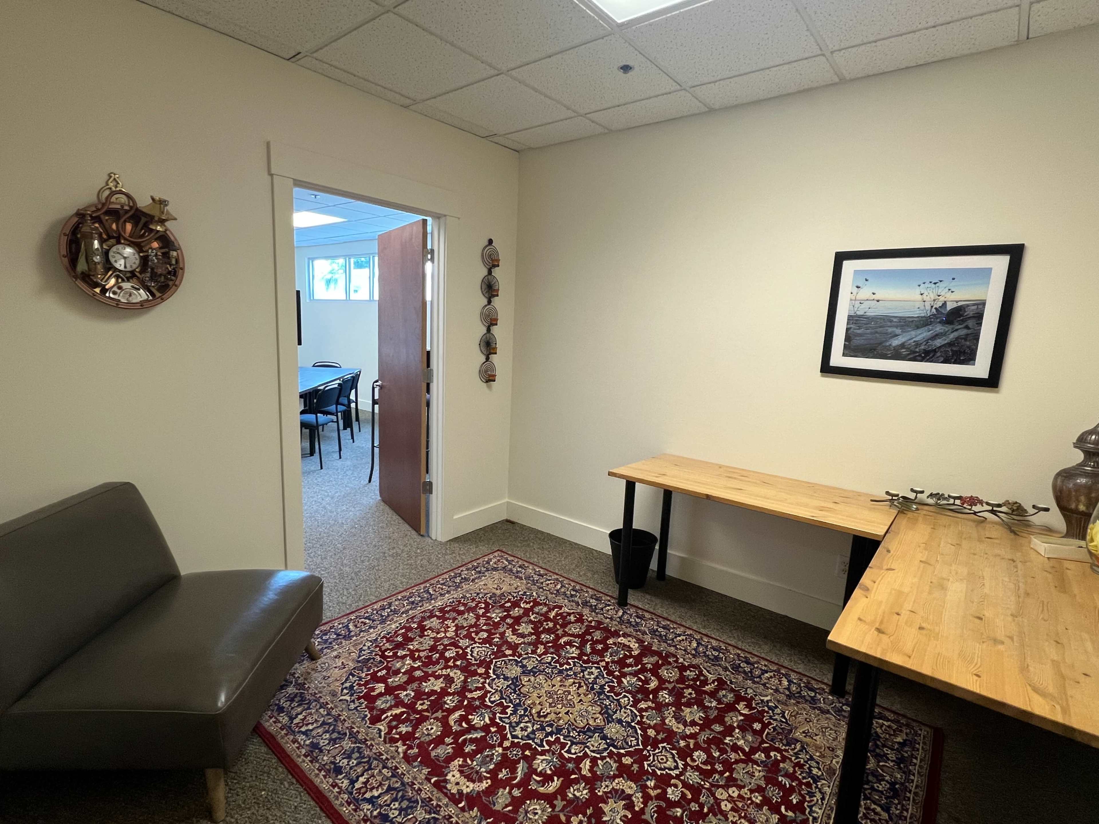 A small office space featuring a cozy seating area, a wooden table, a decorative rug, and an open door leading to a meeting room.