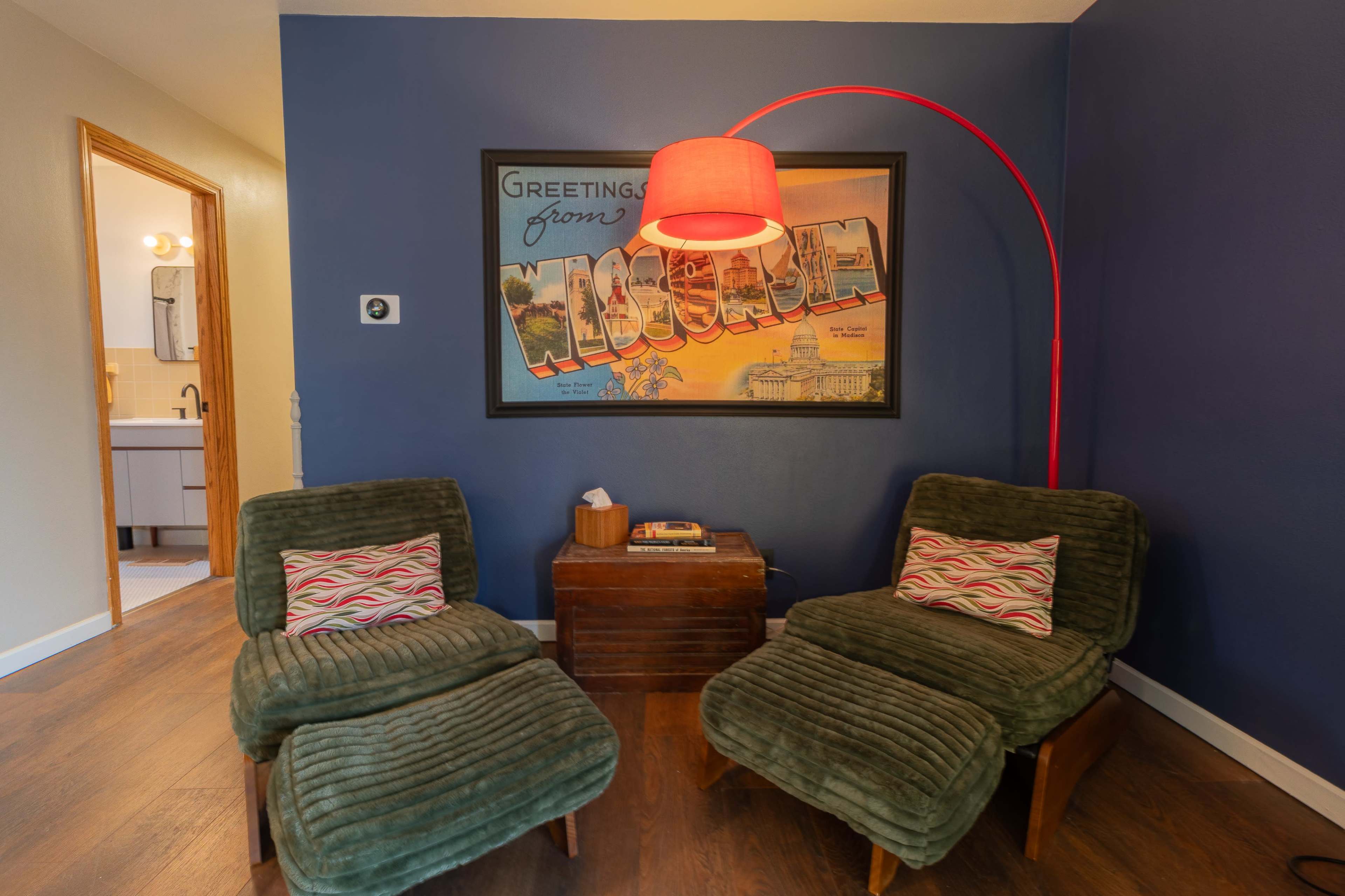 Two green upholstered chairs with a colorful throw pillow are positioned beside a wooden table, beneath a red arc lamp, in a room featuring a vintage "Greetings from Moscow" poster on the wall.