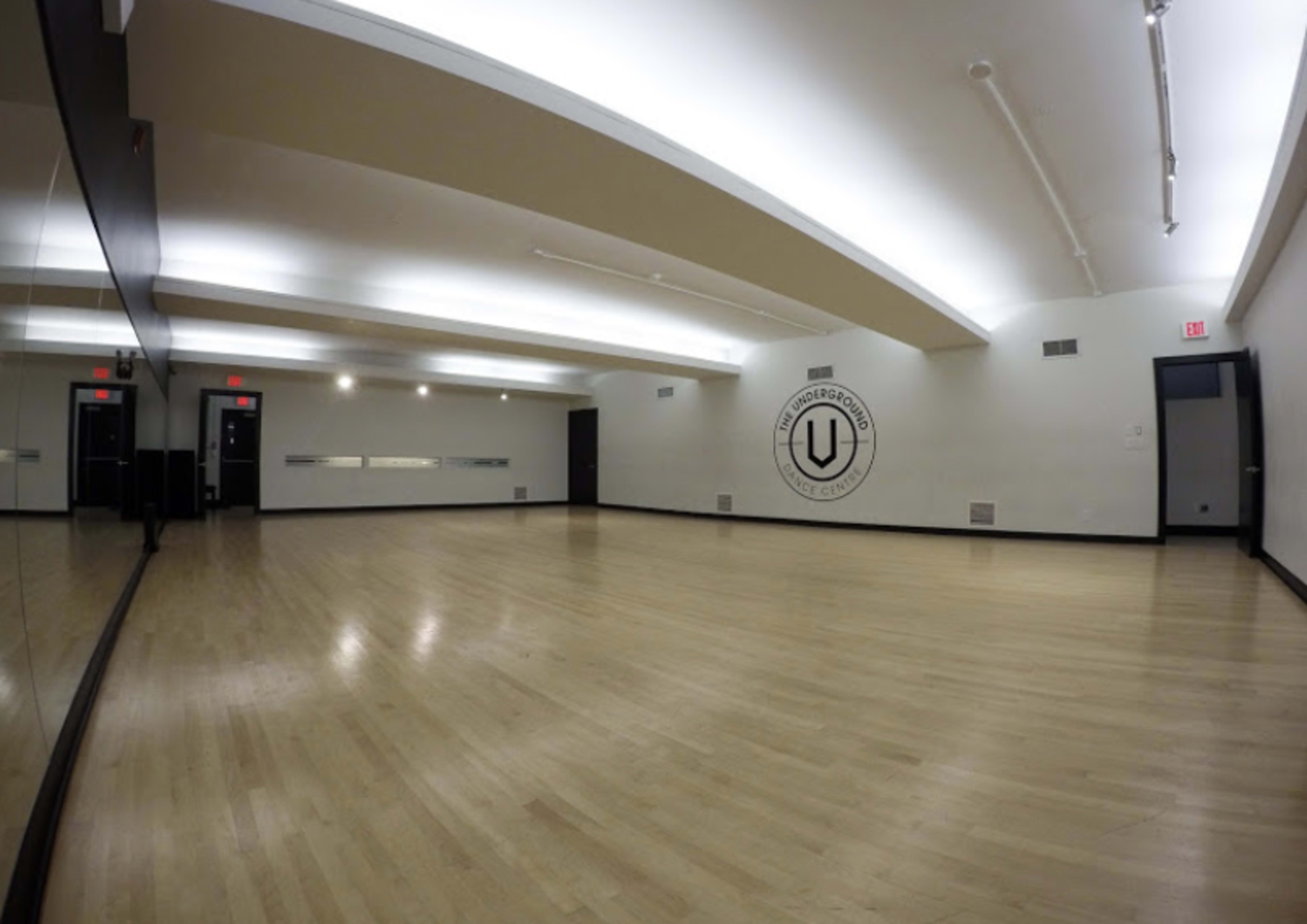 A spacious dance studio with wooden floors, mirrored walls, and a logo on the back wall.