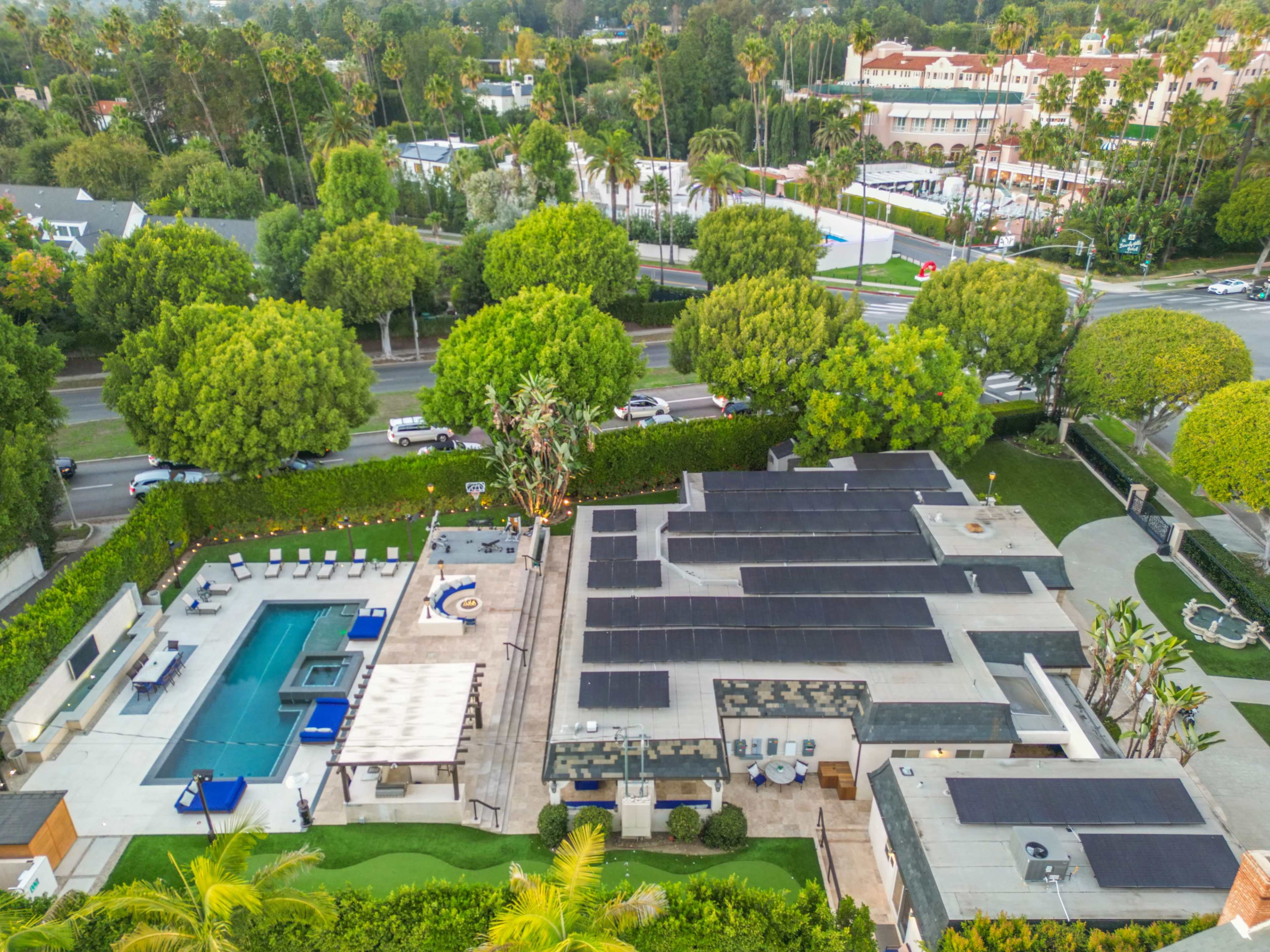 The image shows a modern residential property with a swimming pool, solar panels on the roof, and a landscaped yard surrounded by trees.