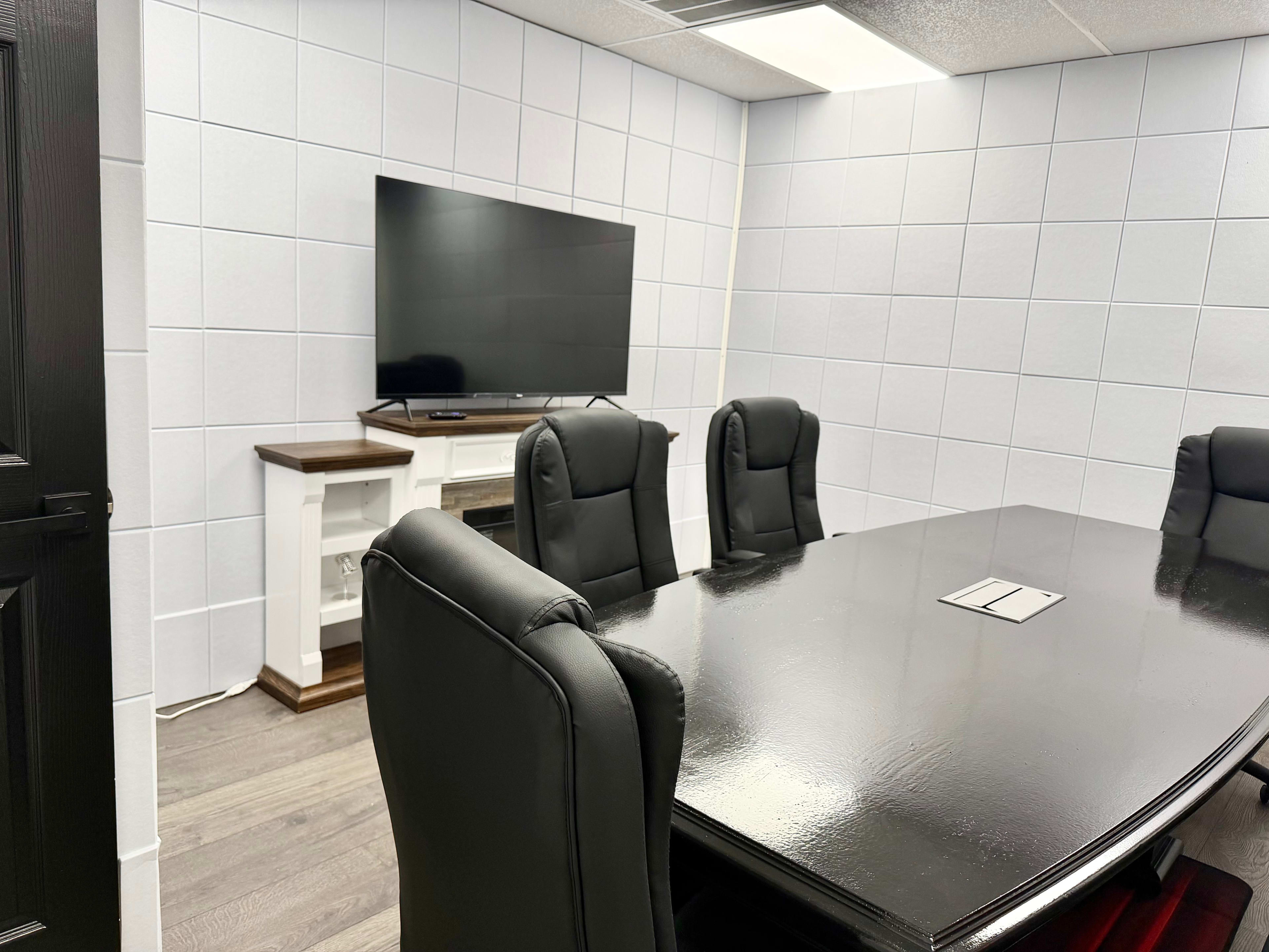 The image shows a conference room with a large black table, black leather chairs, a wall-mounted television, and white paneling.